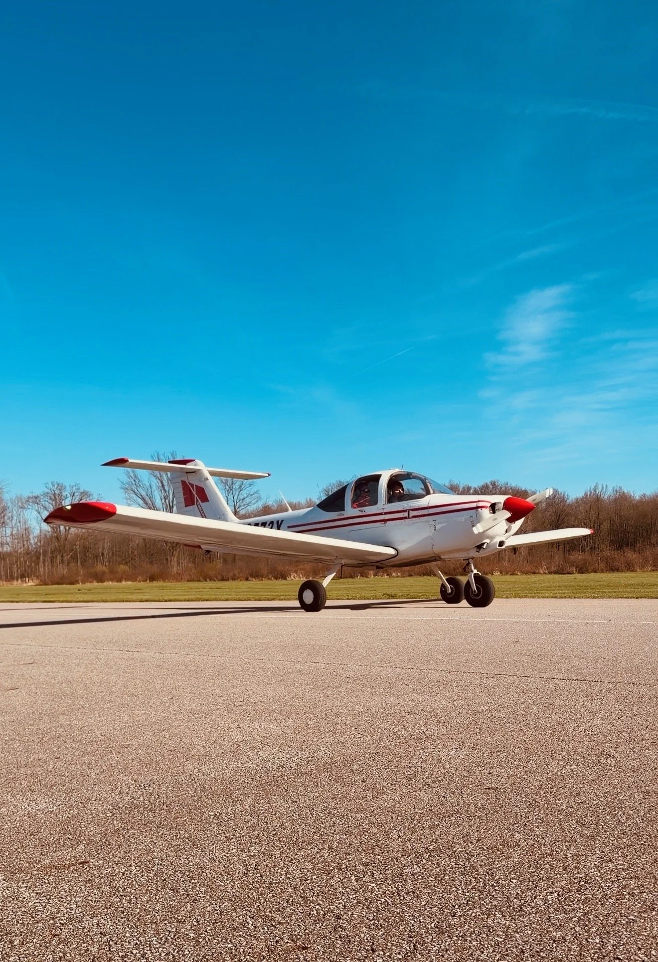 A small white and red propeller airplane on a runway, with trees and a blue sky in the background.