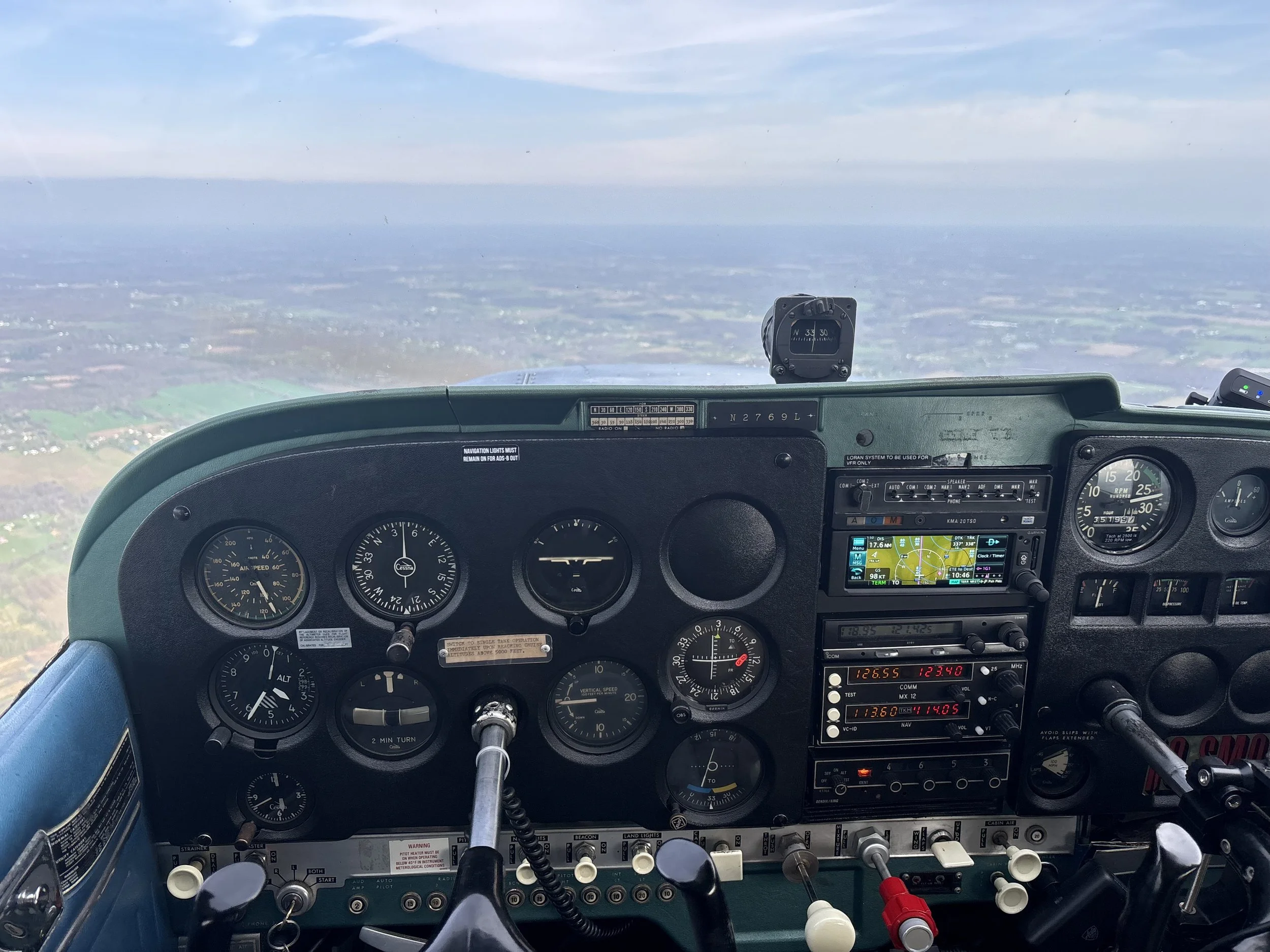 The cockpit of a small airplane showing the instrument panel with gauges, switches, and navigation systems. Through the windshield, a clear sky and a landscape of fields are visible.