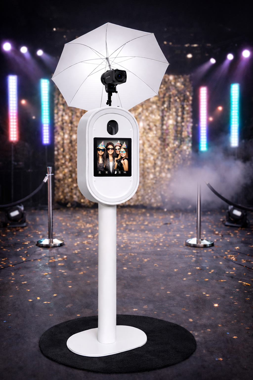 A photo booth with a camera on top and a white umbrella, displaying a screen with three people wearing party hats and sunglasses, at a party or celebration event with colorful lights and a gold backdrop.