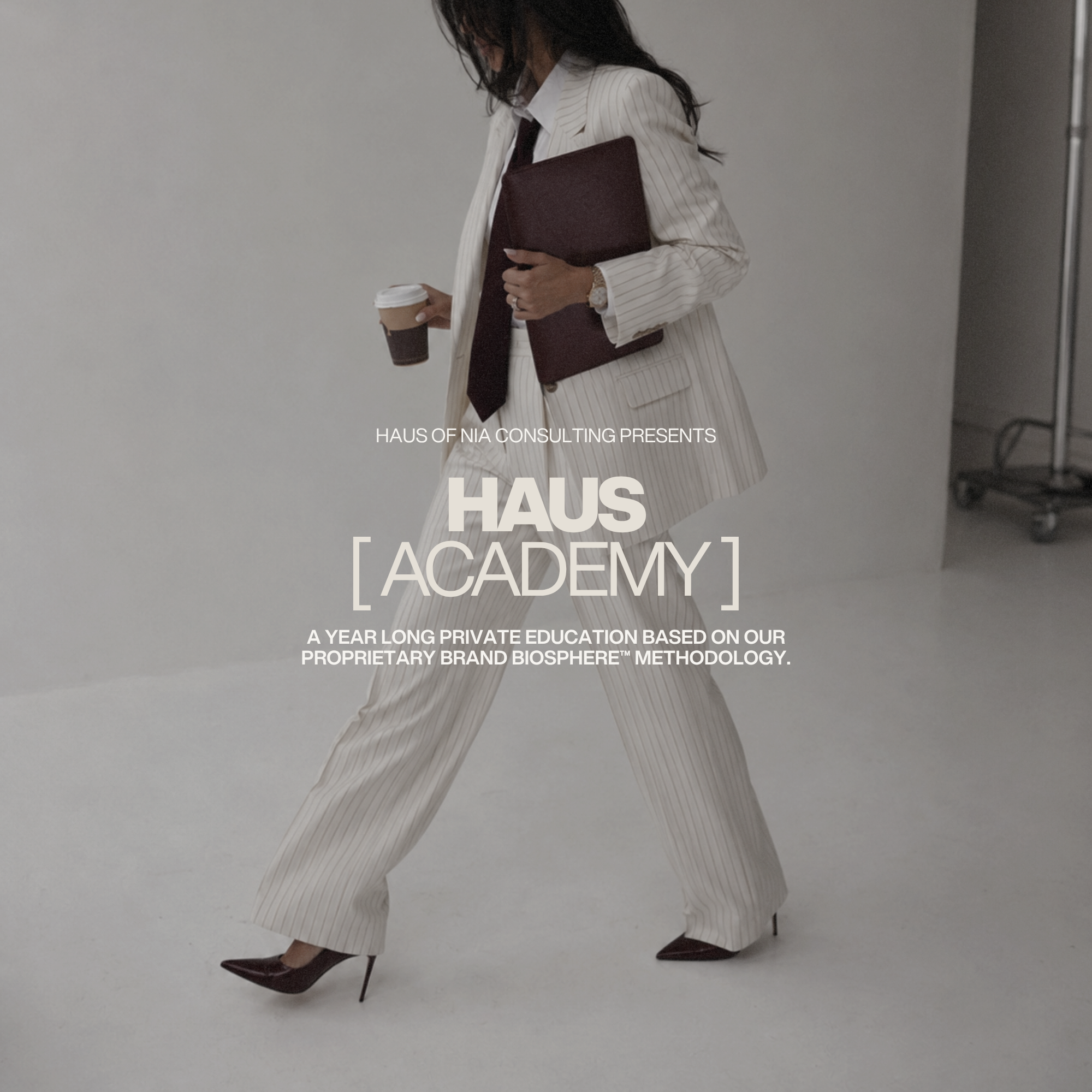 Nia, Founder & Managing Partner, Haus of Nia Consulting Brand Photoshoot - Nia (a woman) dressed in a white pinstripe suit with a black tie, holding a cup of coffee and a maroon folder, standing against a plain gray background.