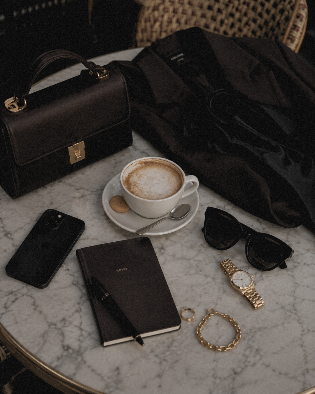 A marble table with a black handbag, a black notebook with a pen, a smartphone, a pair of sunglasses, a gold wristwatch, a gold bracelet, a ring, and a cup of coffee with a cookie. Some black clothing or a bag is partially visible in the background.