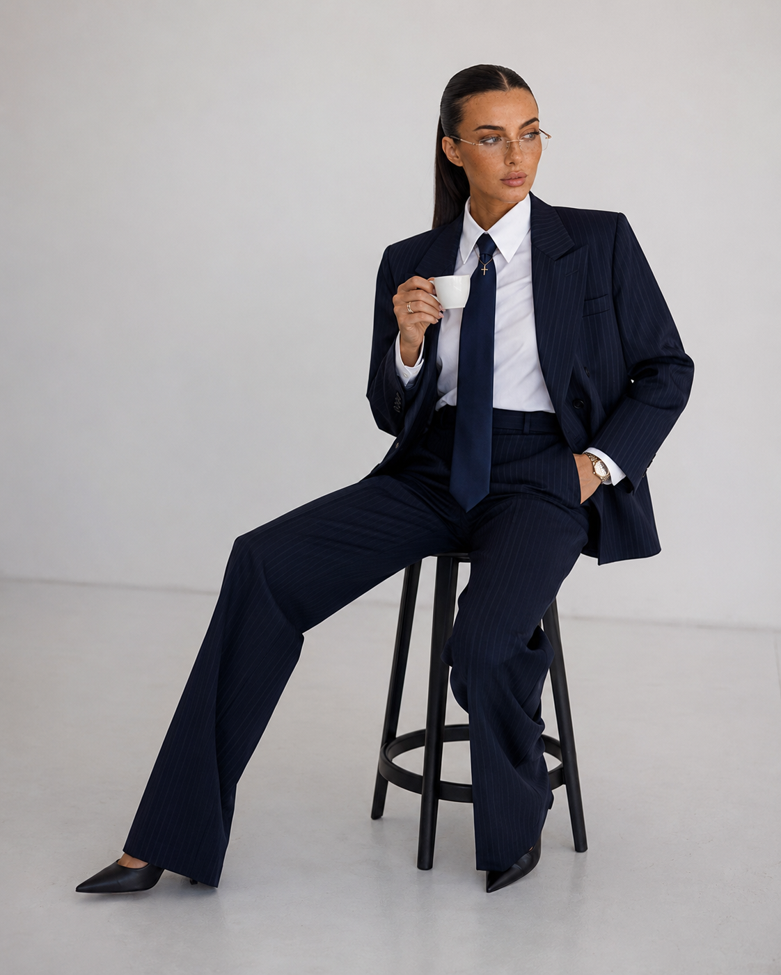 A woman in a navy pinstripe suit sitting on a black stool, holding a white coffee cup, with one hand in her pocket and looking to the side.
