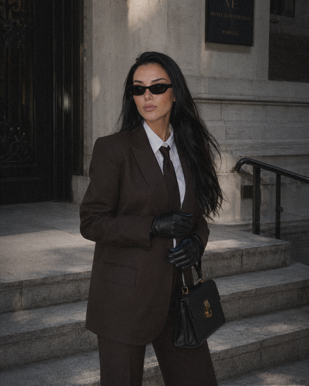 A woman dressed in a business suit with a white shirt and dark tie, wearing sunglasses and black leather gloves, holding a black handbag, standing on steps outside a stone building with a sign that reads 'Hotel & Residences'.