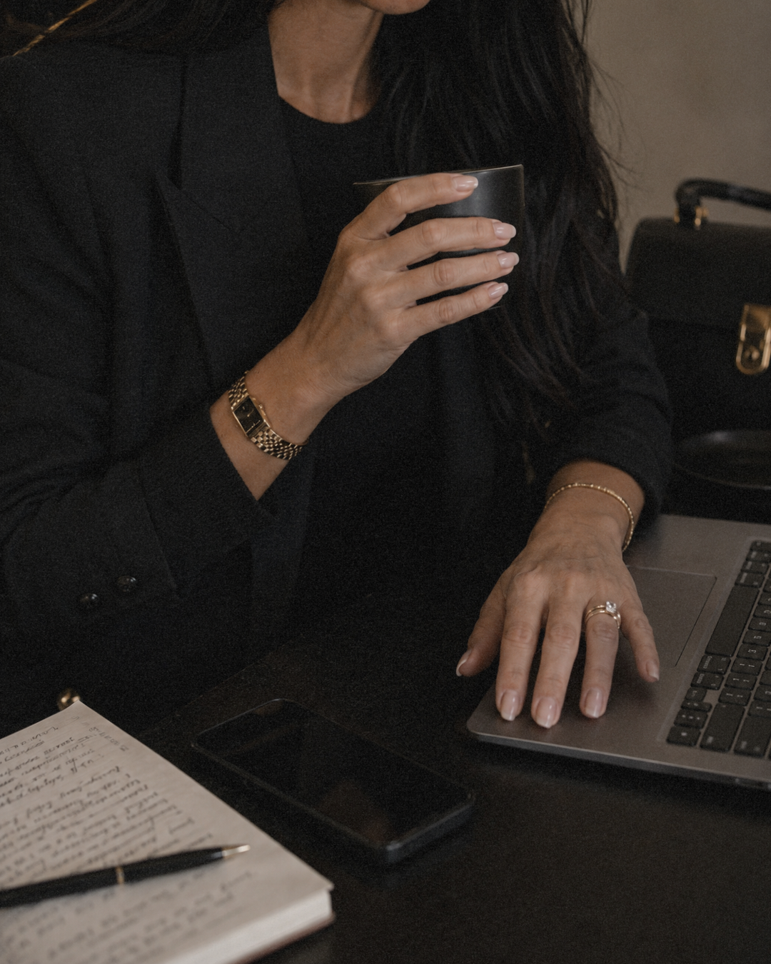 A woman in a black jacket holding a black mug with her right hand, working on a laptop at a desk. The desk has a notebook with handwritten notes, a pen, a smartphone, and a black handbag in the background.