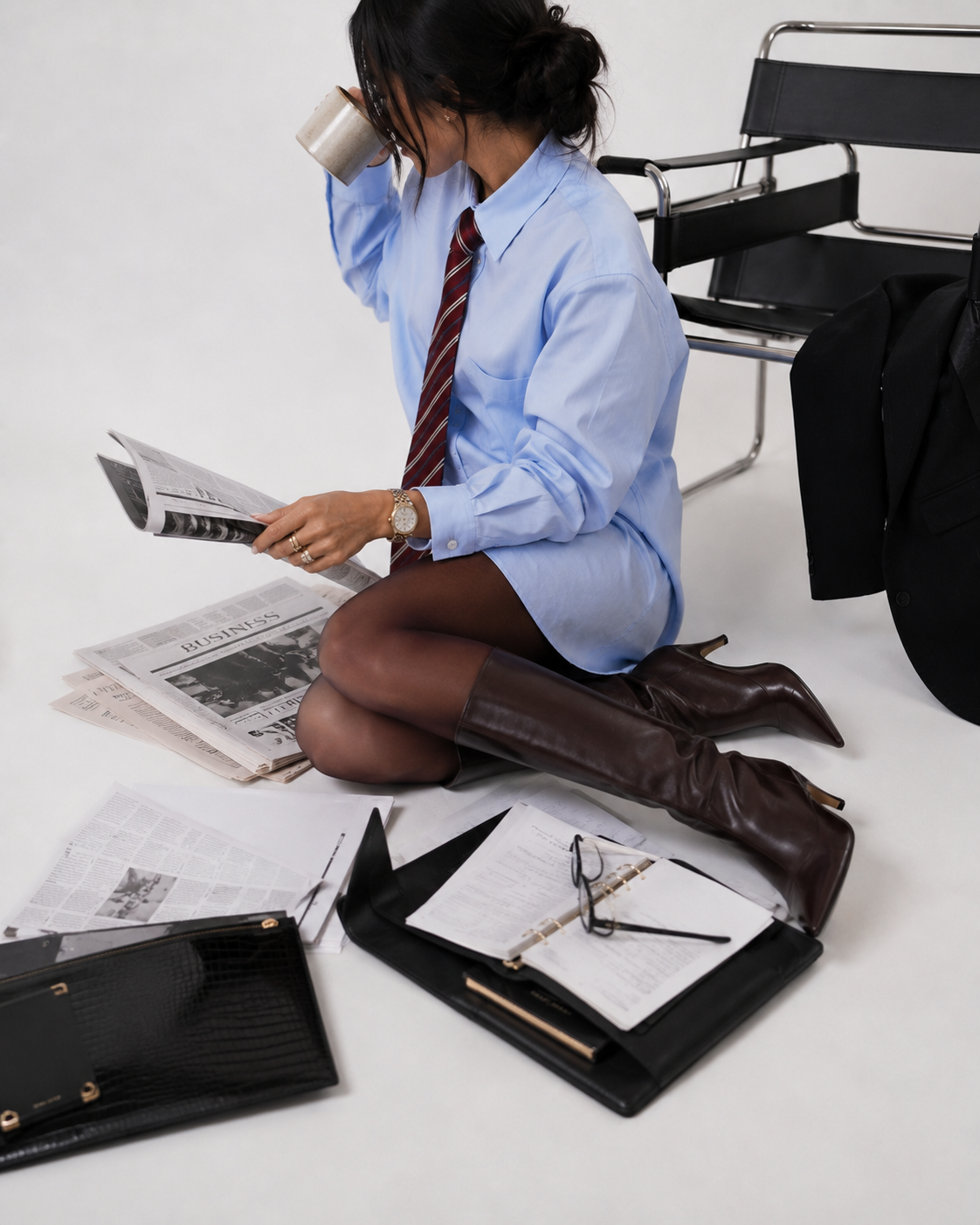 Woman in business attire sitting on the floor, reading a newspaper, surrounded by scattered papers, glasses, and office accessories.