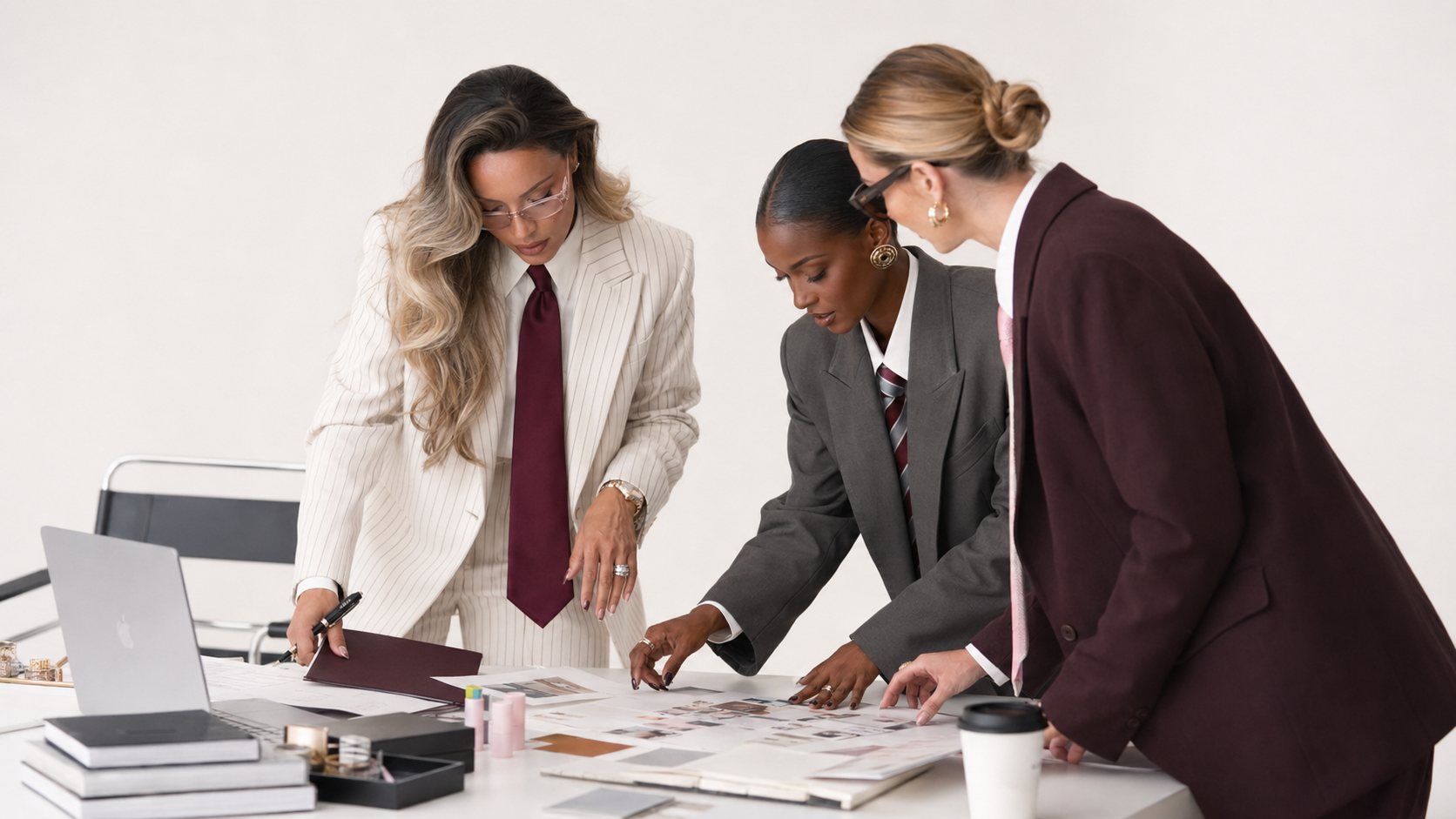 Three women in business suits are gathered around a table, examining documents and images.