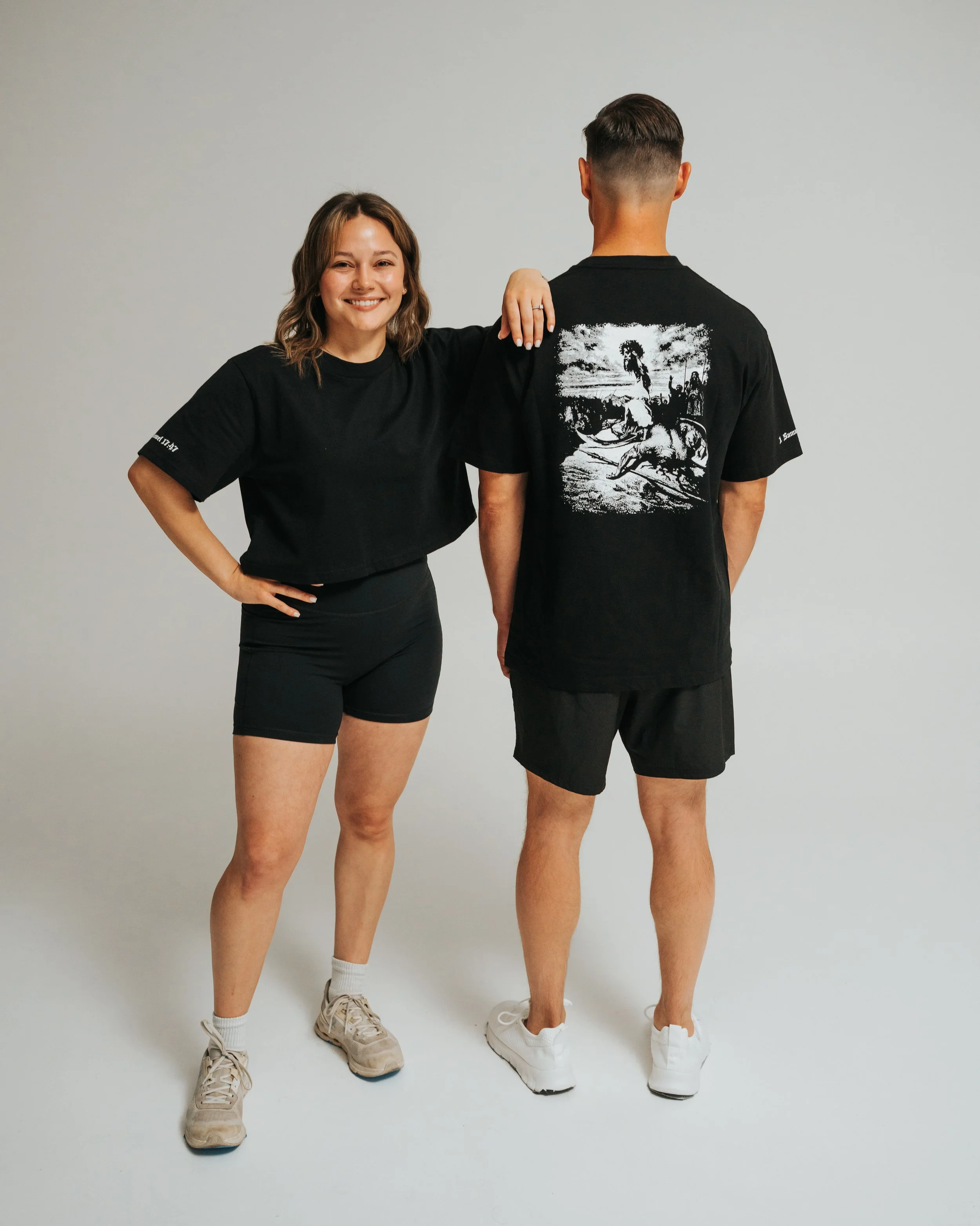 A woman with shoulder-length hair smiles with her hand on her hip, wearing a black t-shirt and shorts, standing next to a man with his back turned, wearing a black graphic t-shirt and shorts, against a plain gray background.