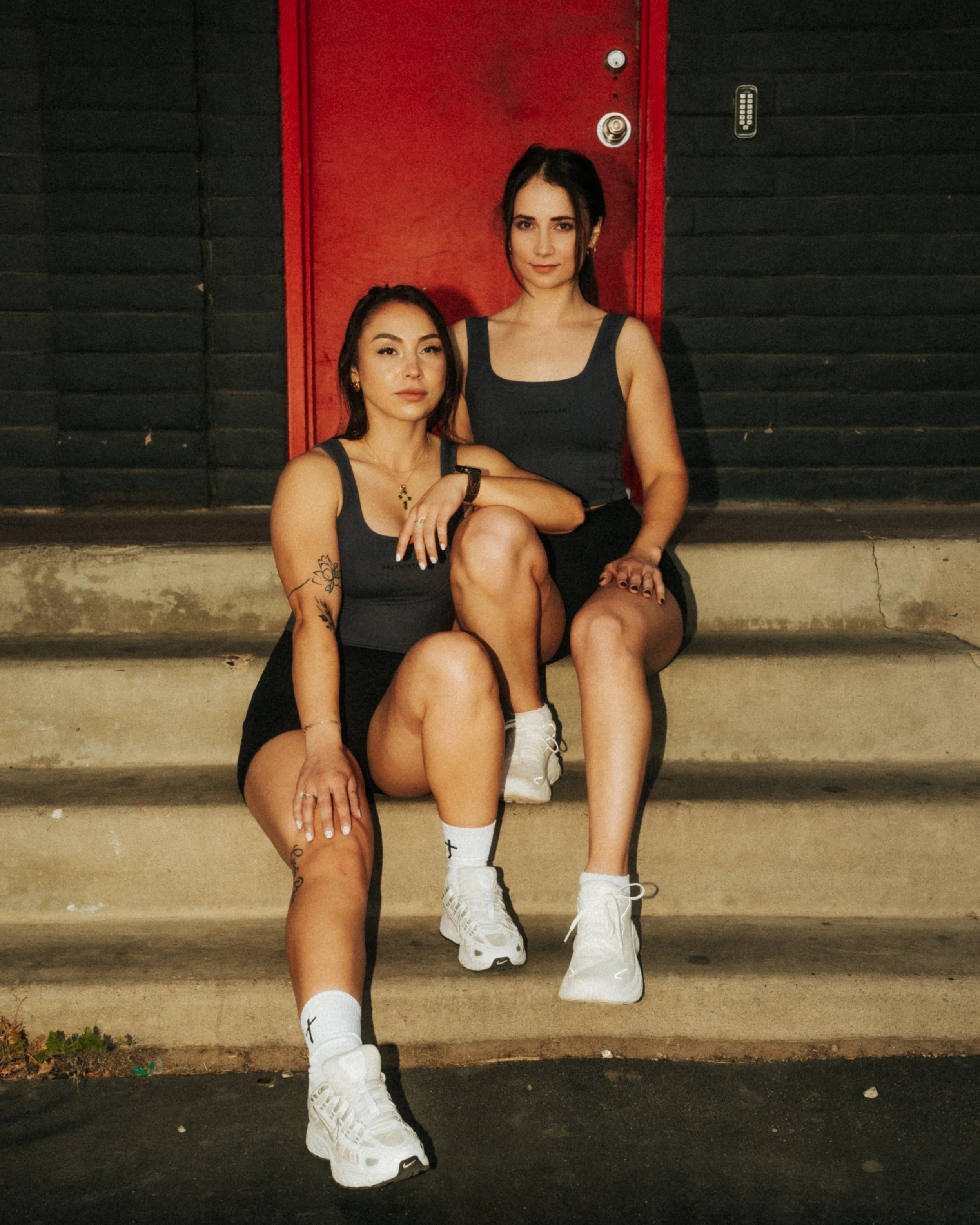 Two young women sitting on concrete steps in front of a red door with black walls, both wearing black tank tops and athletic shoes, with tattoos and jewelry.