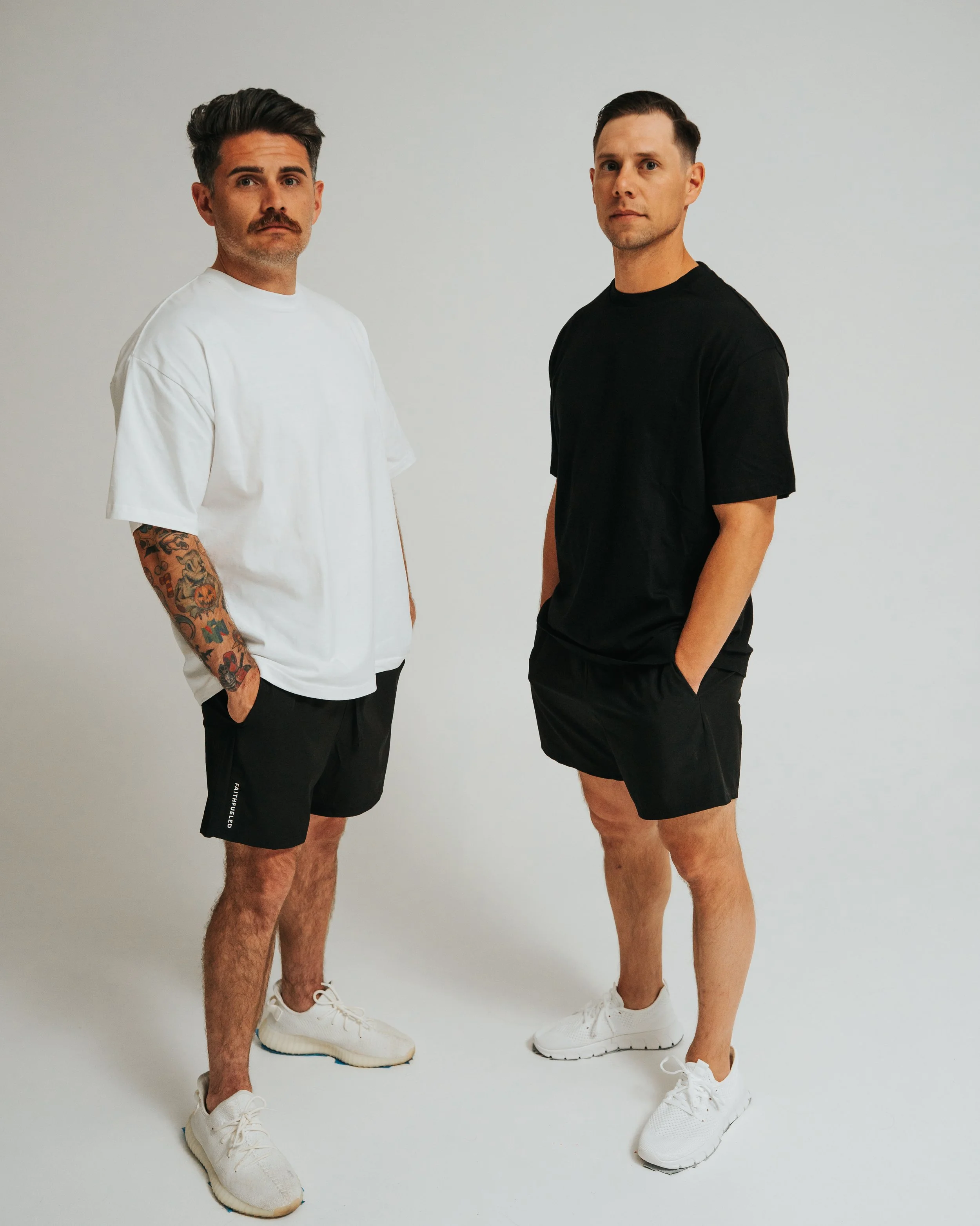 Two men standing side by side against a plain white background, both dressed in casual athletic wear with their hands in their pockets, looking at the camera.