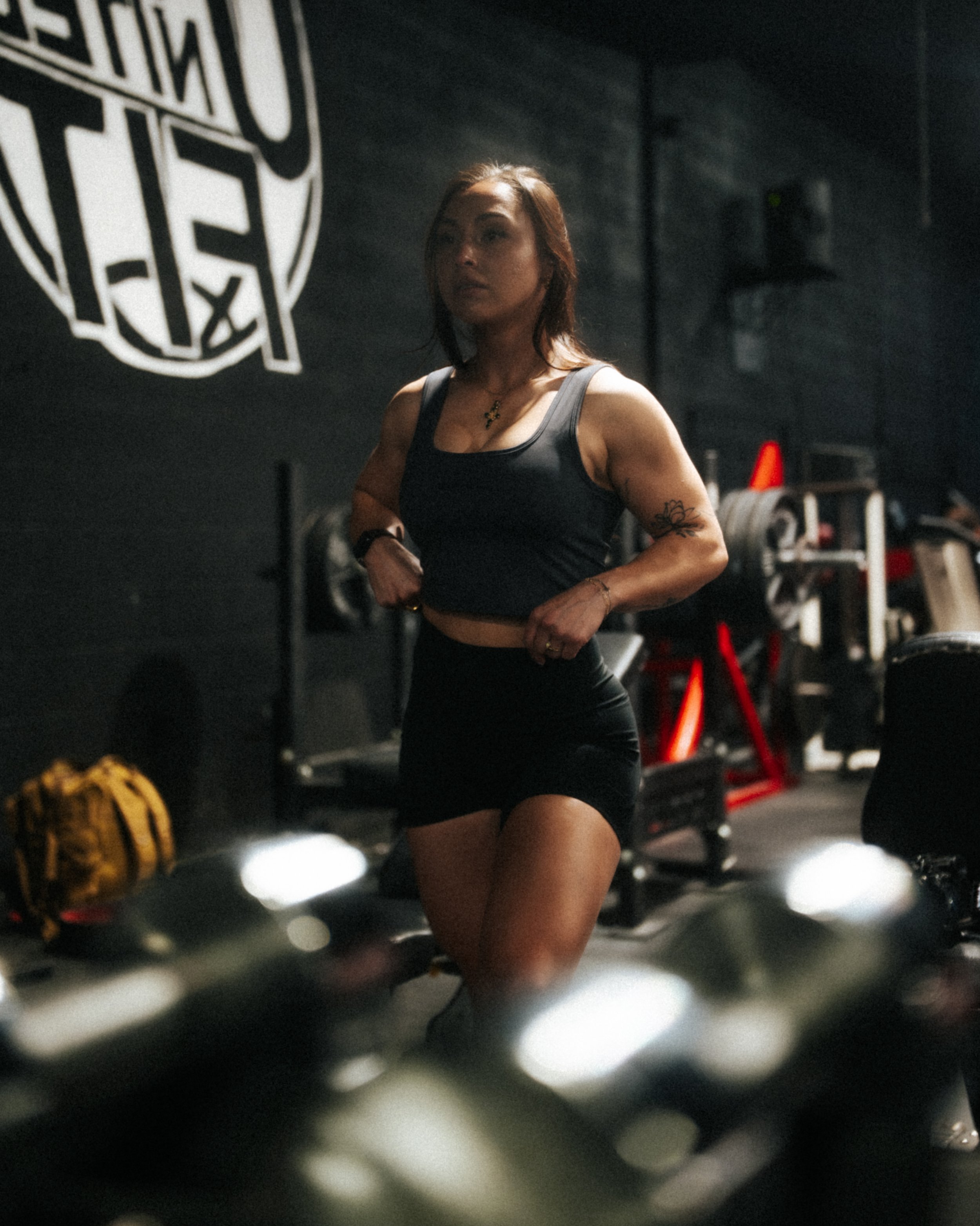 A woman with reddish-brown hair, wearing a black sports bra and black shorts, running on a treadmill in a dimly lit gym, with gym equipment and a large black and white logo on the wall in the background.