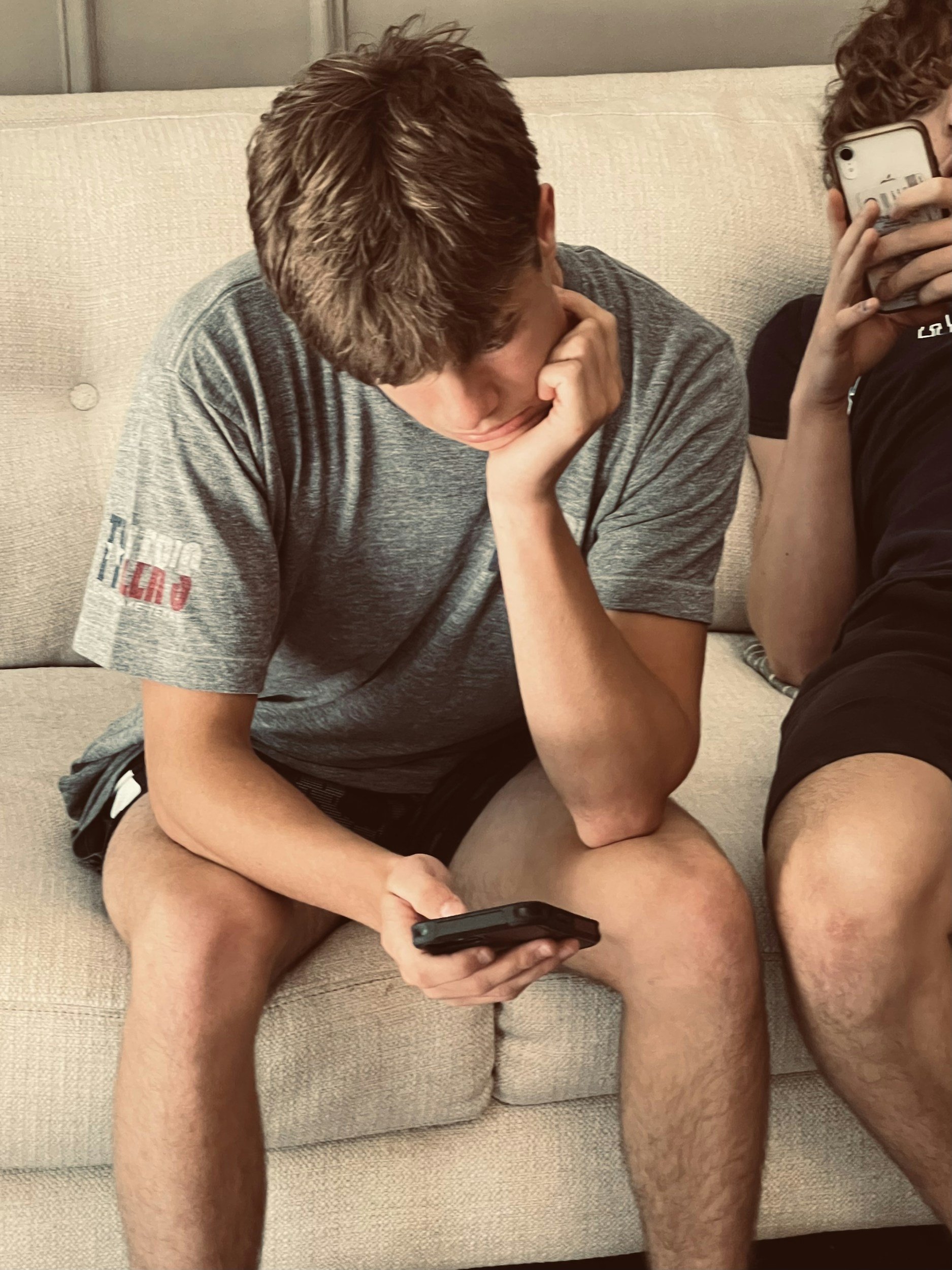 Two teenage boys sitting on a beige couch, both looking at their phones. The boy on the left has brown hair and wears a gray T-shirt and black shorts. The boy on the right has curly hair and is partially visible, holding a phone in front of his face.