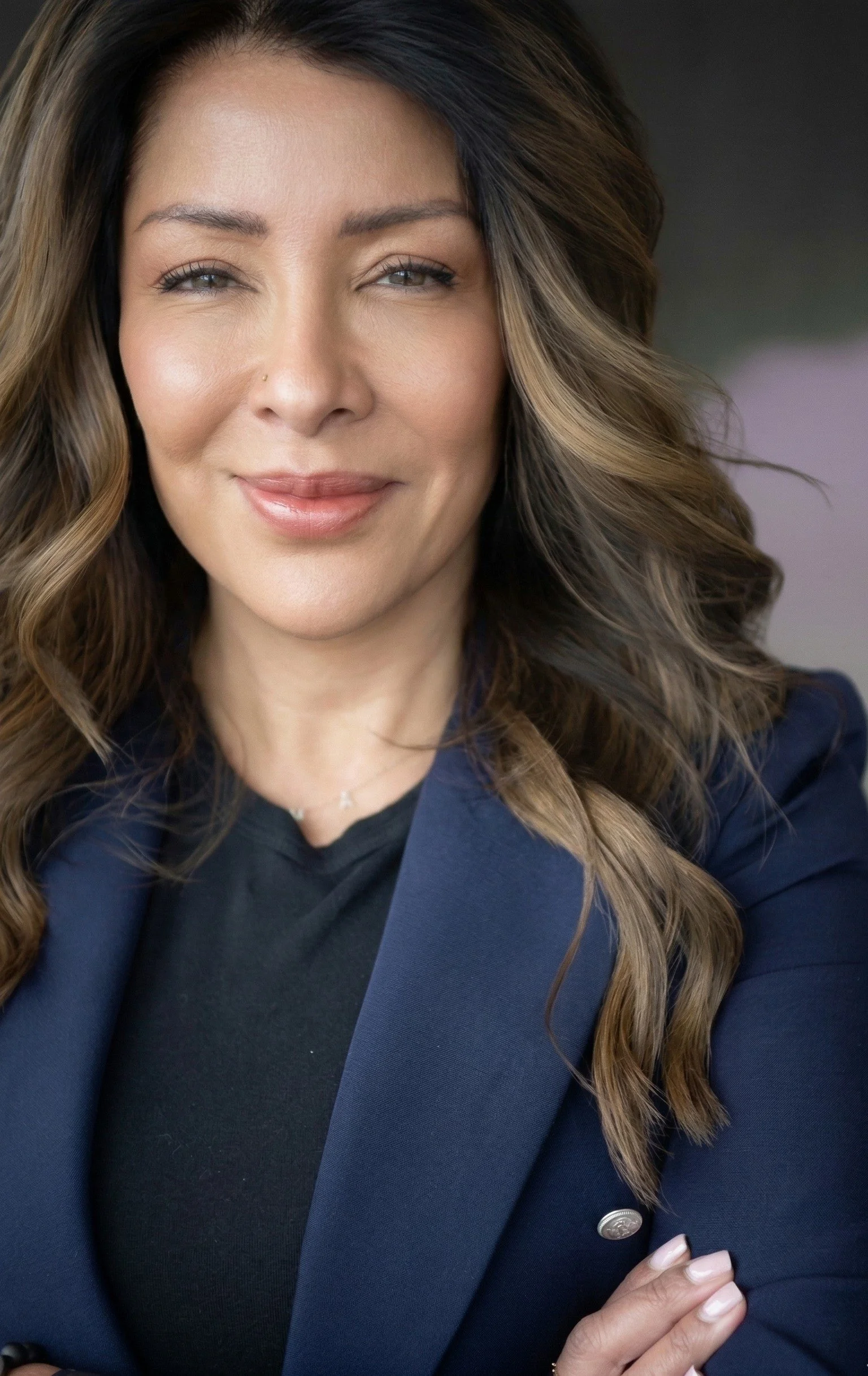 Close-up of a woman with long wavy brown hair, wearing a navy blue blazer and black top, smiling softly, with a blurred background.