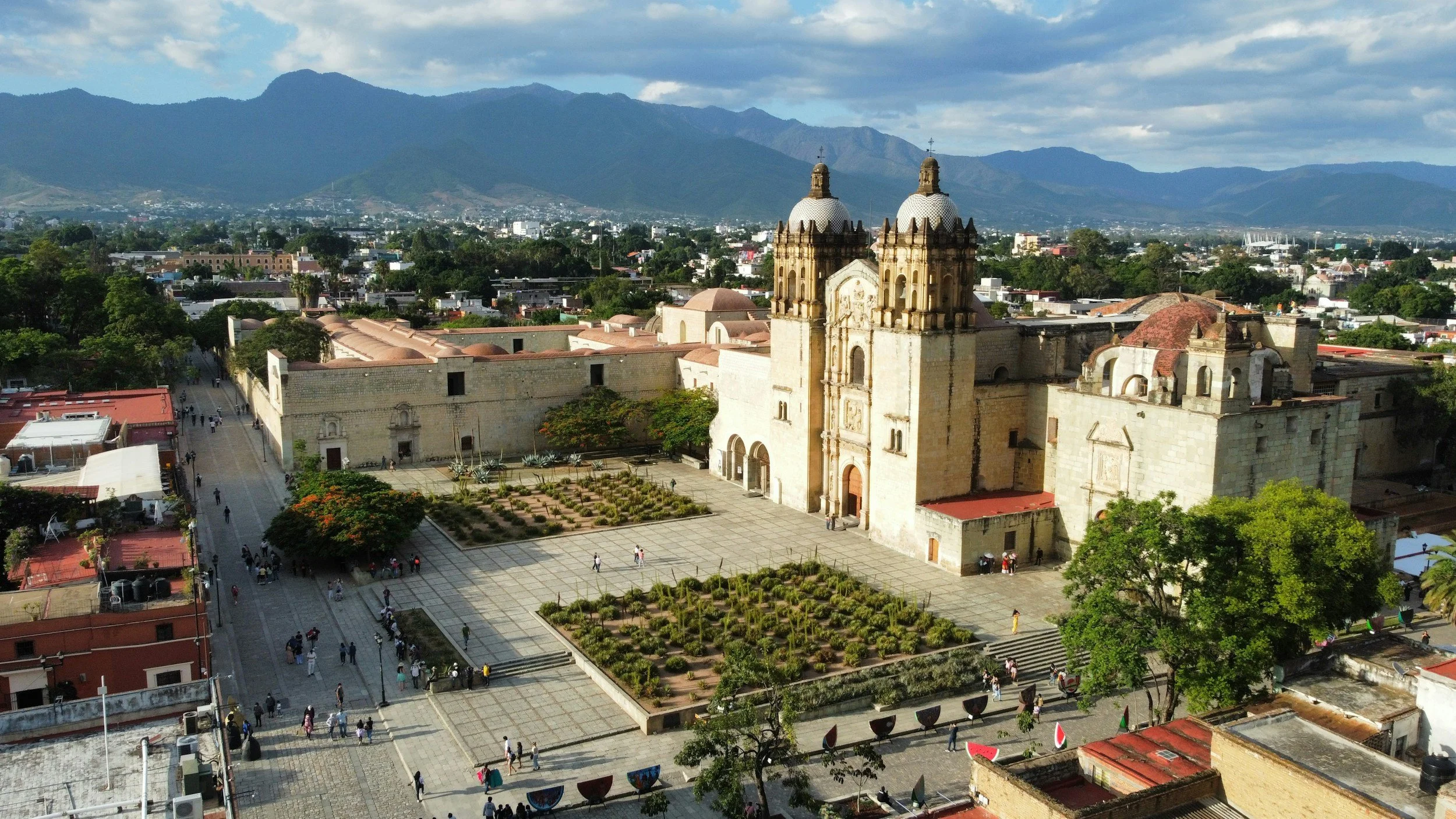 An aerial view of a historic church with twin towers in a city with mountains in the background. The church is surrounded by a plaza with small gardens and people walking.