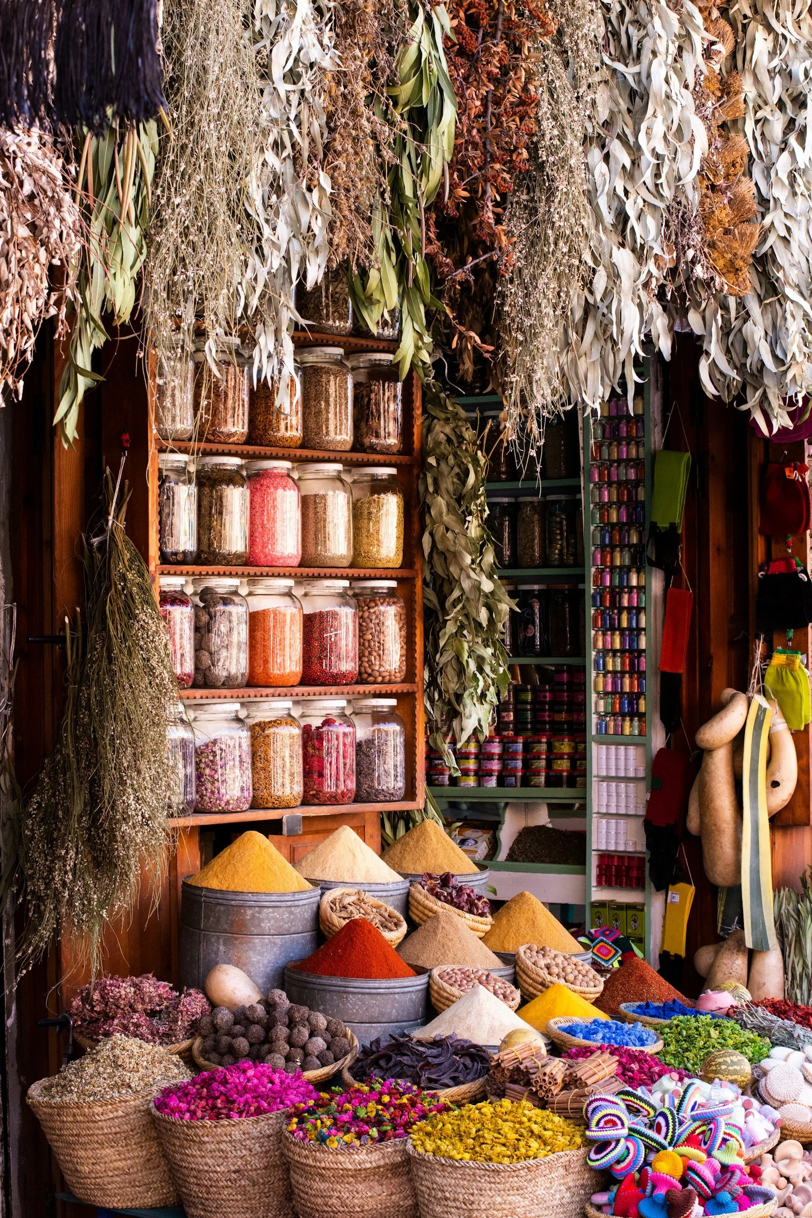 Display of dried herbs, spices, and colorful trinkets at a market stall with jars of spices on shelves behind, hanging herbs, and baskets of flowers and spices in front.