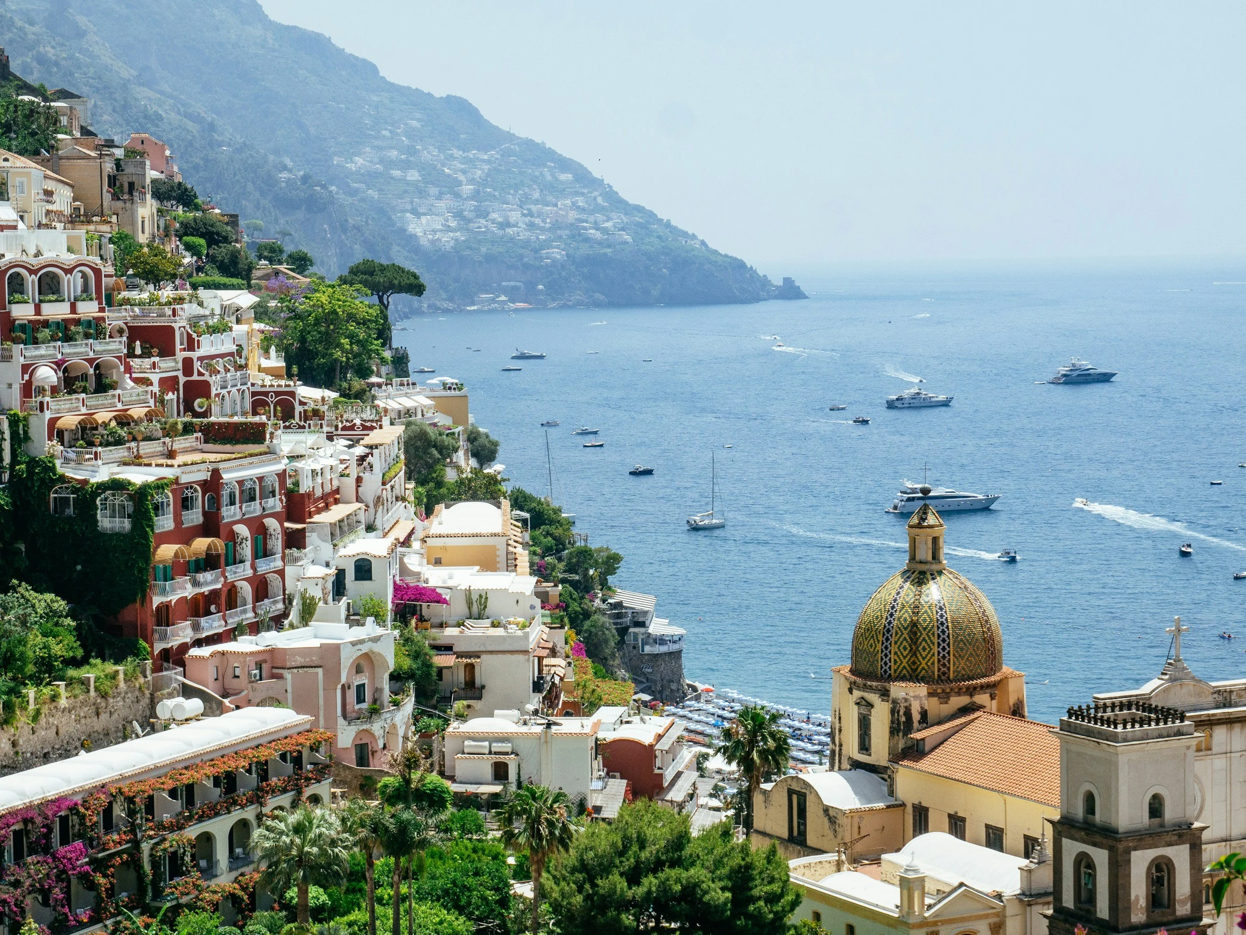 Colorful coastal town with buildings on a hillside overlooking the ocean, with boats in the water and a church with a decorative dome in the foreground.