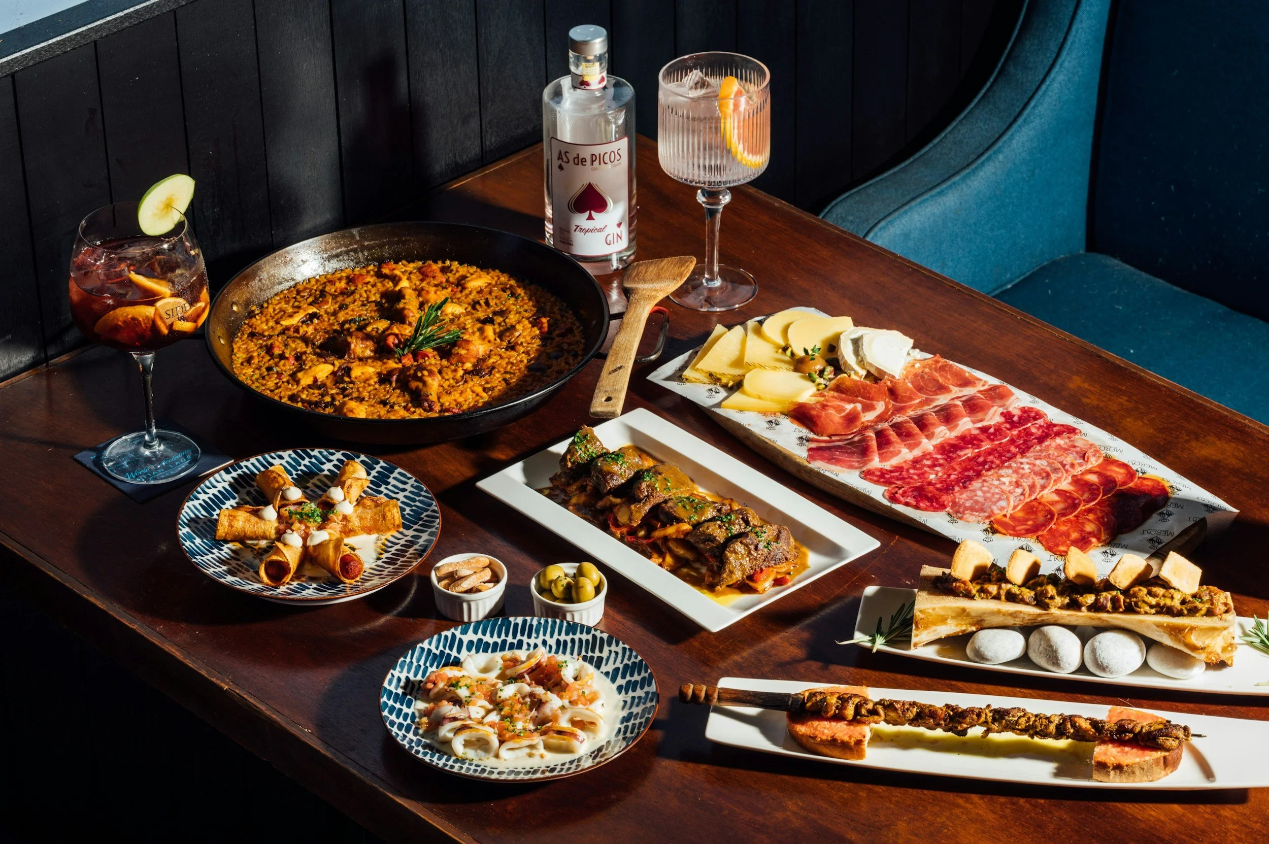 A table with Spanish tapas and drinks, including a large pan of paella, a charcuterie board with cheeses and cured meats, plates of fried appetizers, small bowls of olives, and glasses of cocktails with fruit garnishes.