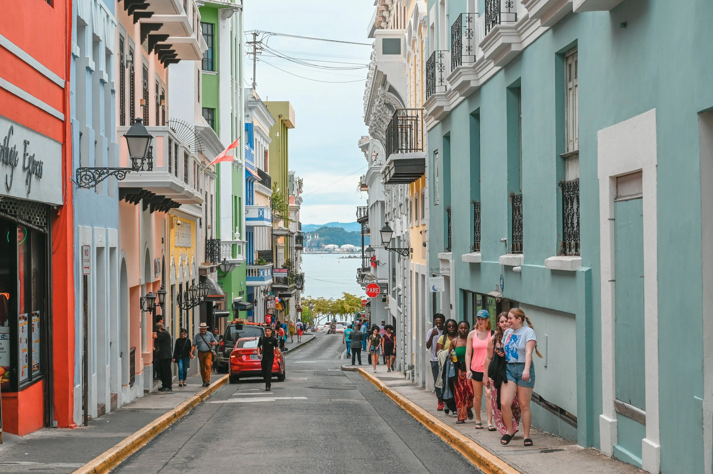 Colorful buildings line a street with pedestrians walking on the sidewalk and cars parked along the curb, overlooking a body of water in the background.