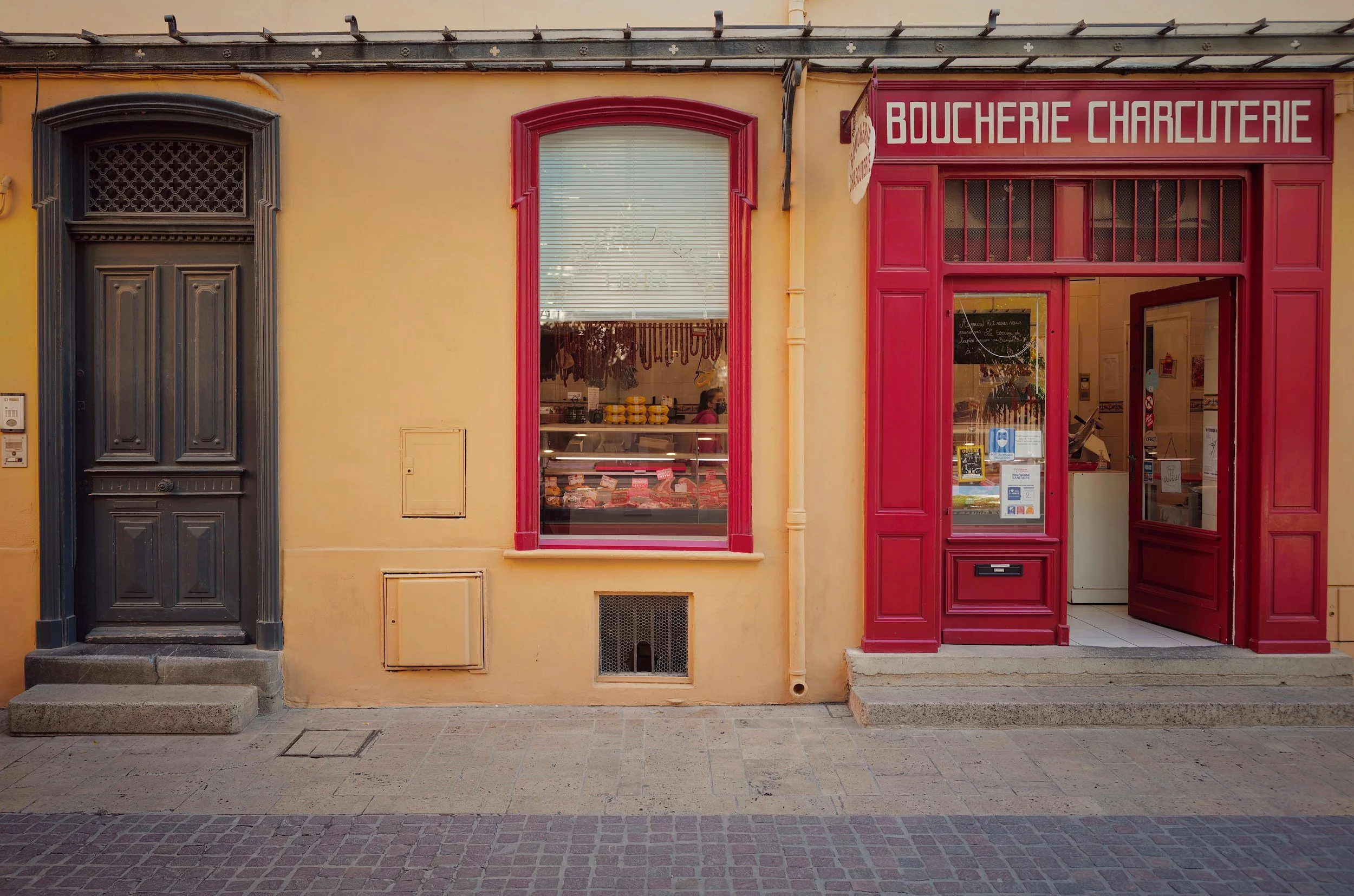 Front of a French butcher shop with a black door on the left and a red-framed window and entrance on the right, featuring a sign that reads 'BOUCHERIE CHARCUTERIE' and visible meat inside.