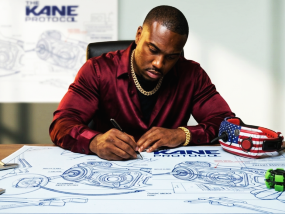 A man in a red satin shirt and gold jewelry signs engineering blueprints on a desk with a remote control with an American flag design and a green watch visible.