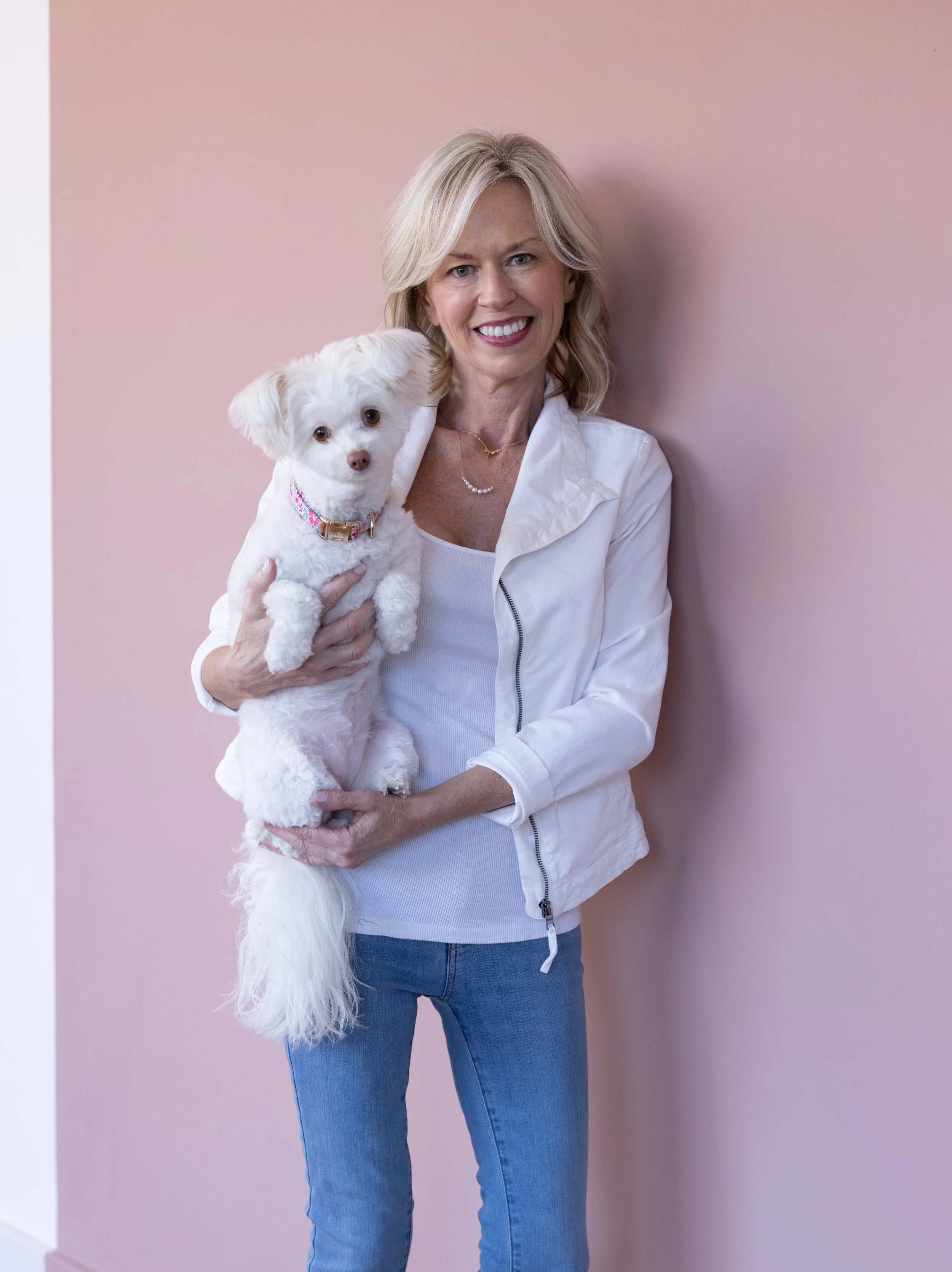 Rachel Doescher smiling and holding her white dog, Noodle, in front of a soft pink background