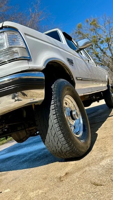 White OBS Ford pickup truck parked on a concrete driveway with trees and a blue sky in the background.
