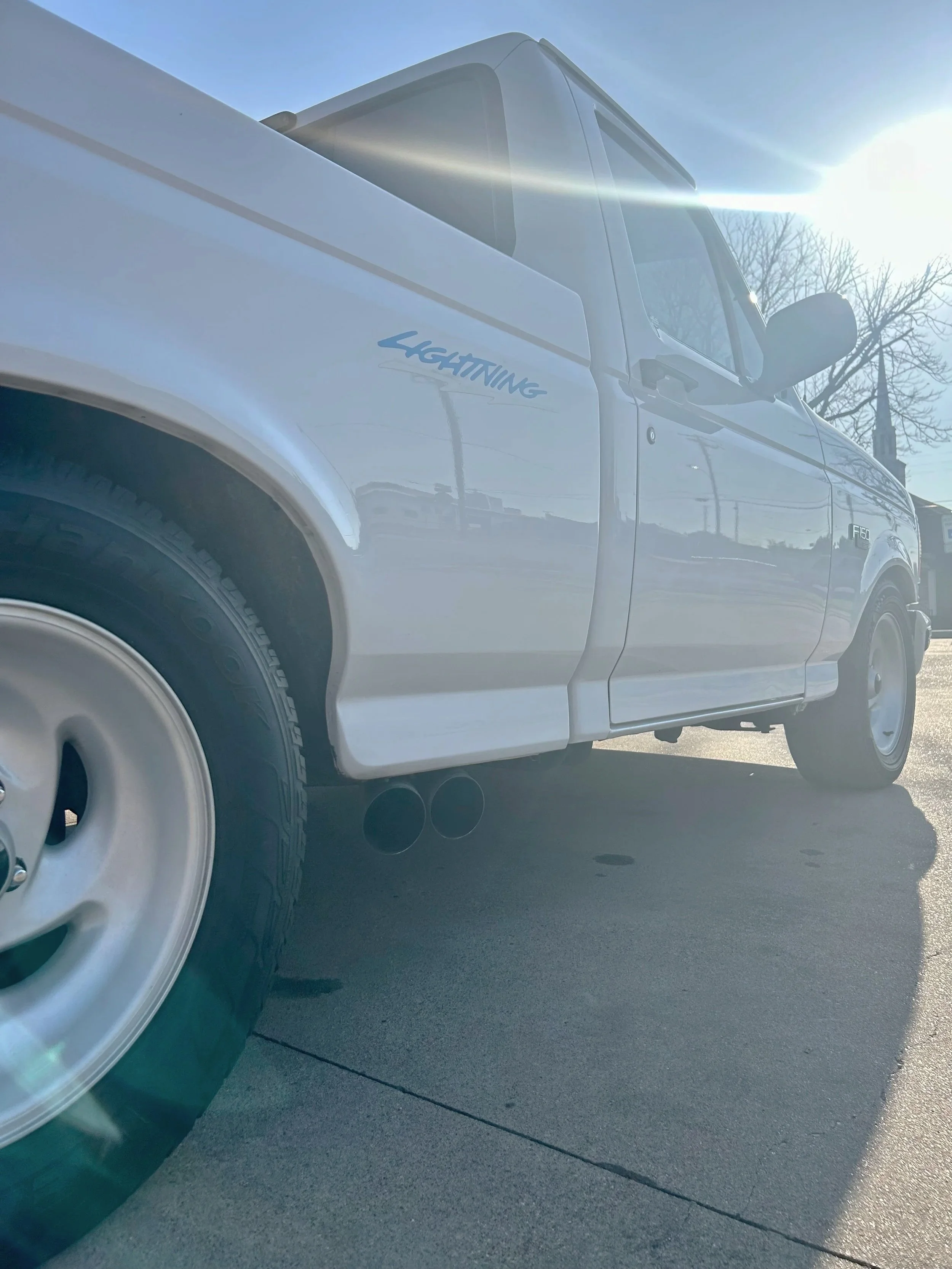 Close-up of a white pickup truck with a 'Lightning' decal parked on a paved surface, with the sun shining brightly in the sky.