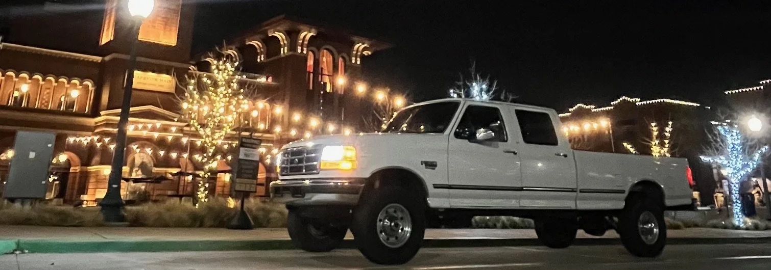 White extended cab pickup truck parked on a city street at night, with decorated buildings and trees illuminated in the background.
