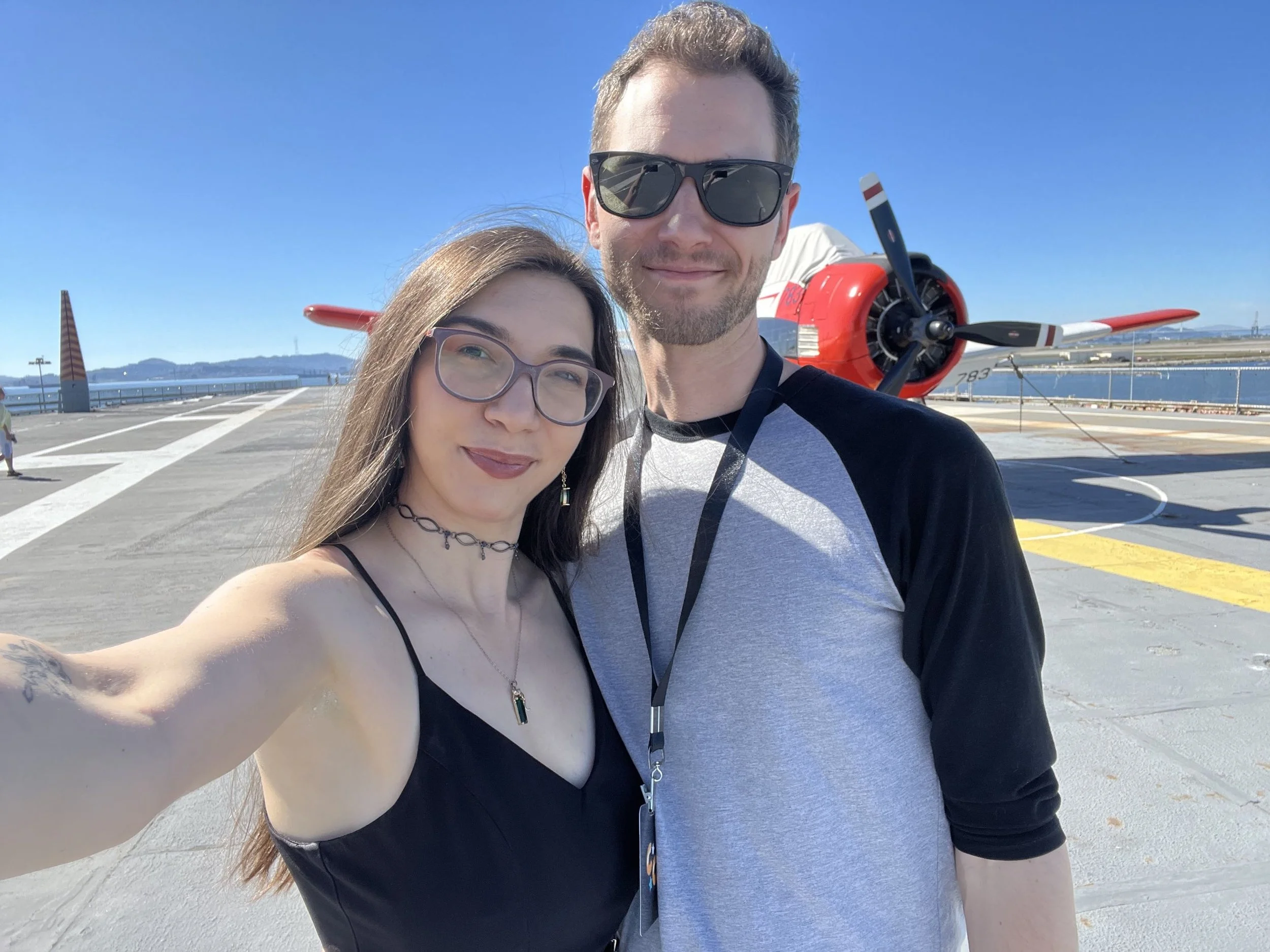 A young woman and a young man are taking a selfie on the deck of an aircraft carrier, with a blue sky and ocean in the background. The woman has long hair, glasses, and is wearing a black top and jewelry. The man has short hair, sunglasses, and is wearing a gray and black baseball tee. There is a red and white propeller airplane in the background.