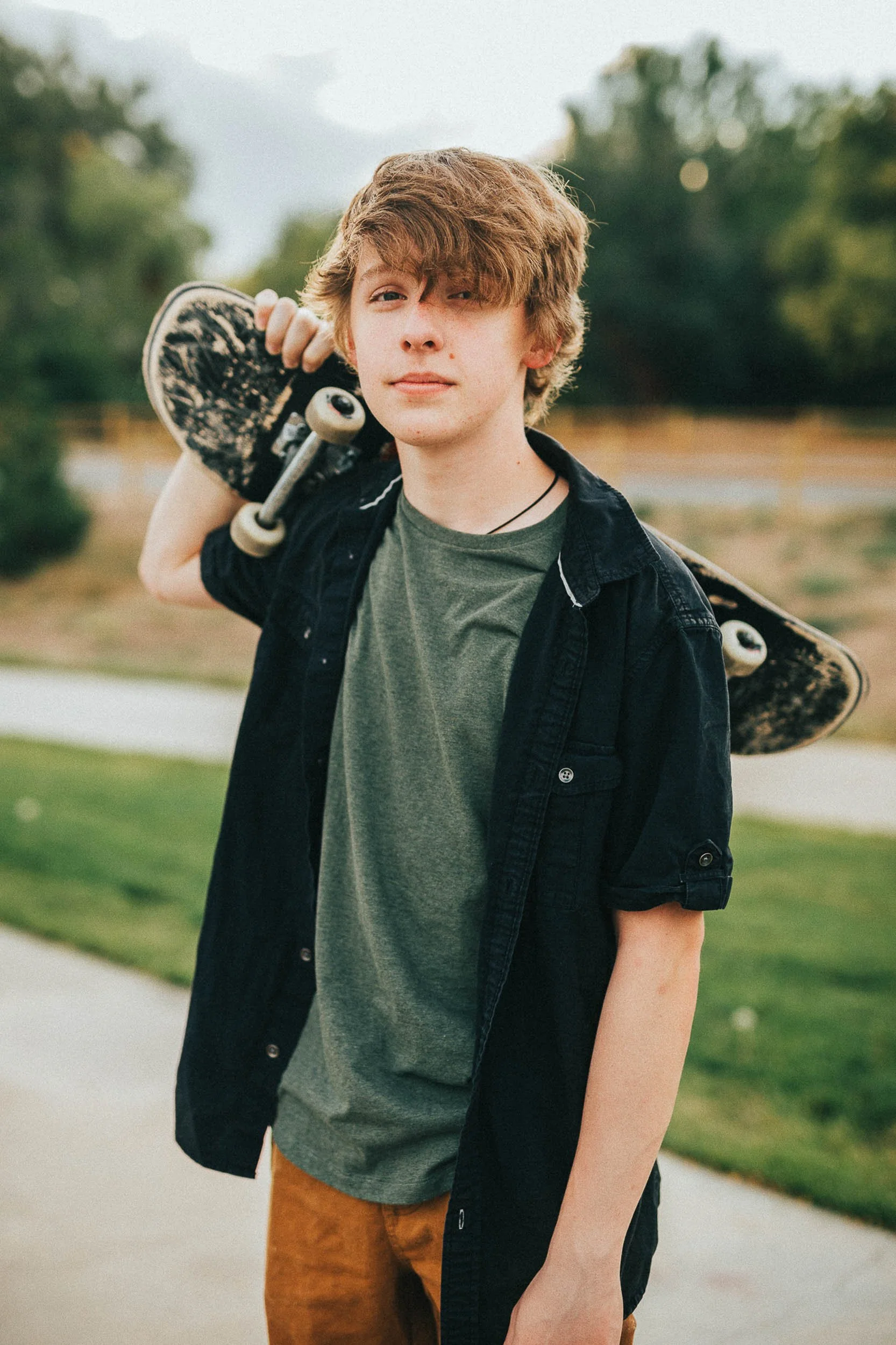 Caucasian high school teen standing in a park with a skateboard over his shoulder posing for senior portraits