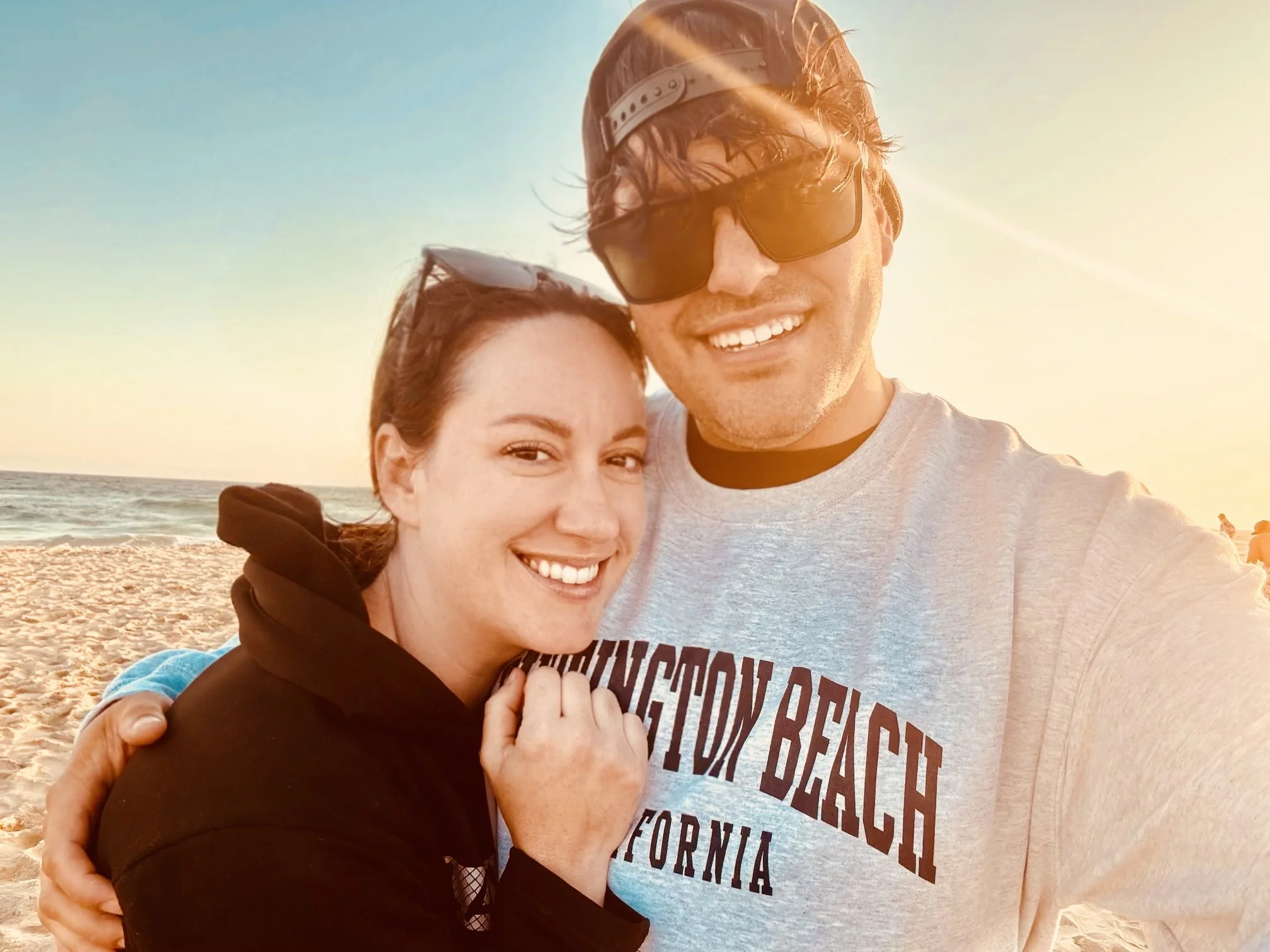 A young woman and a young man smiling at the beach during sunset, standing close together with the woman hugging the man, ocean waves in the background.