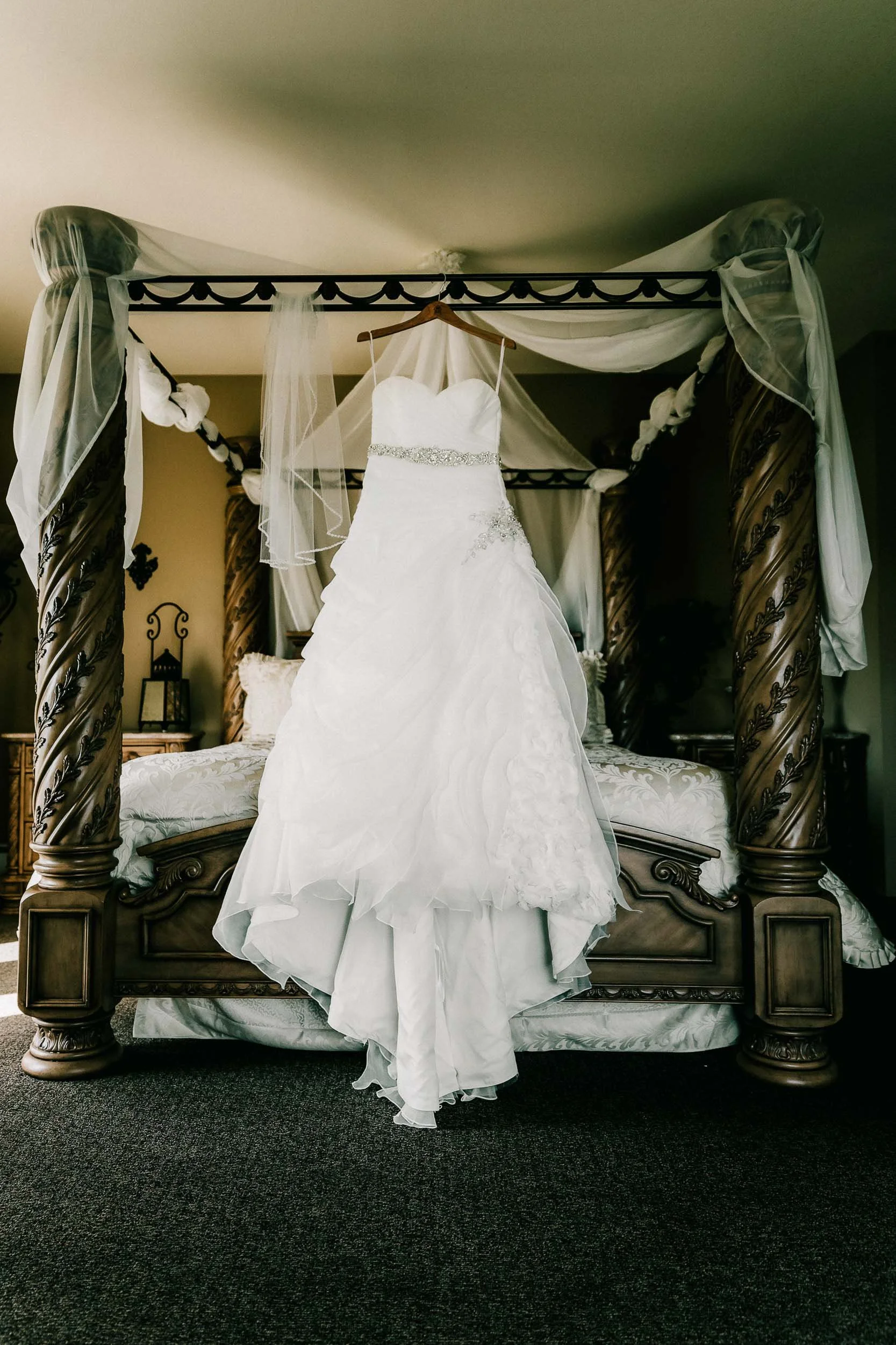 White wedding dress hanging on a wooden hanger from a four-poster bed with dark wood and ornate carvings, in a bedroom with decorative curtains and furnishings.