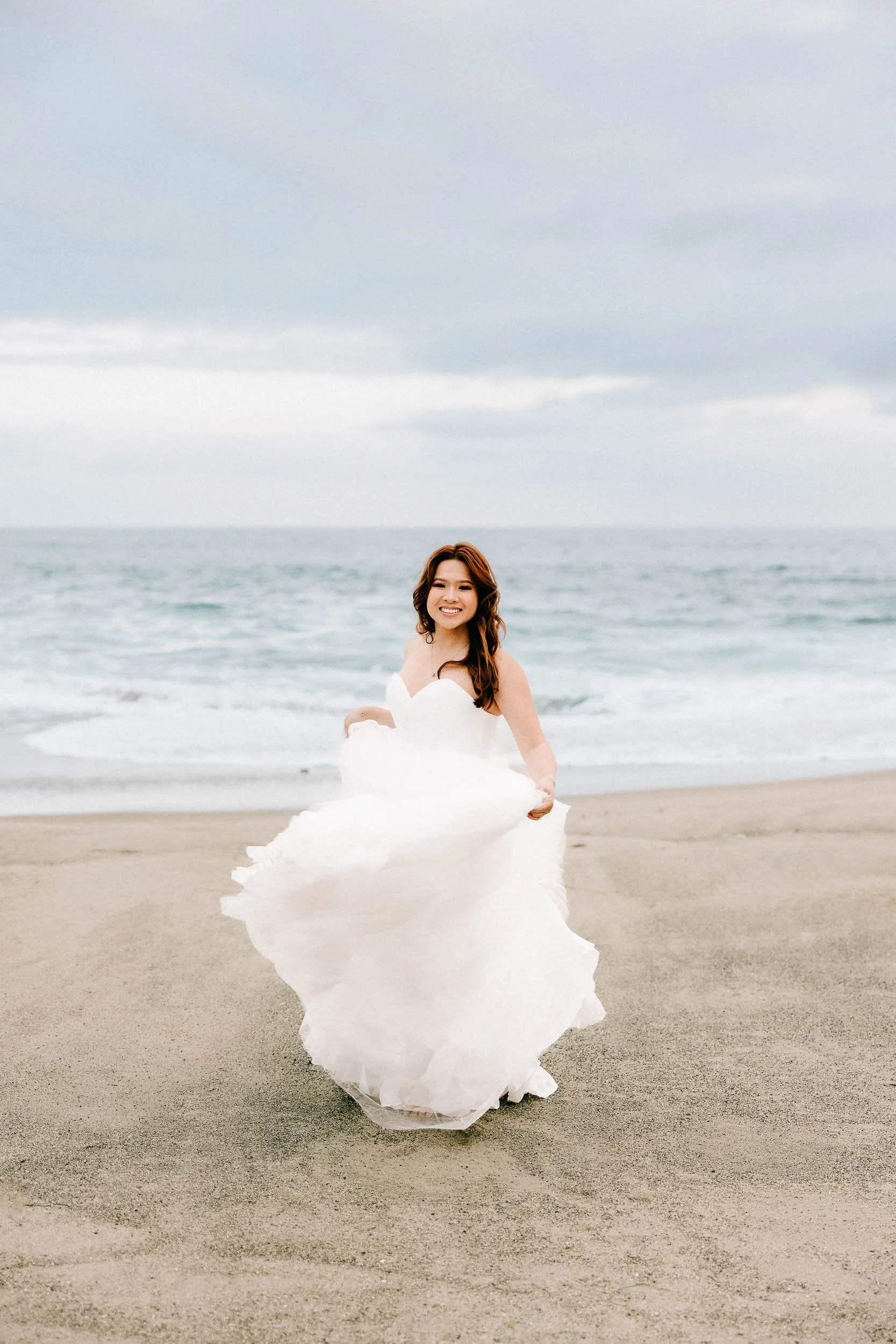 An asian woman in a white wedding dress smiling on a sandy beach with the ocean and cloudy sky in the background.