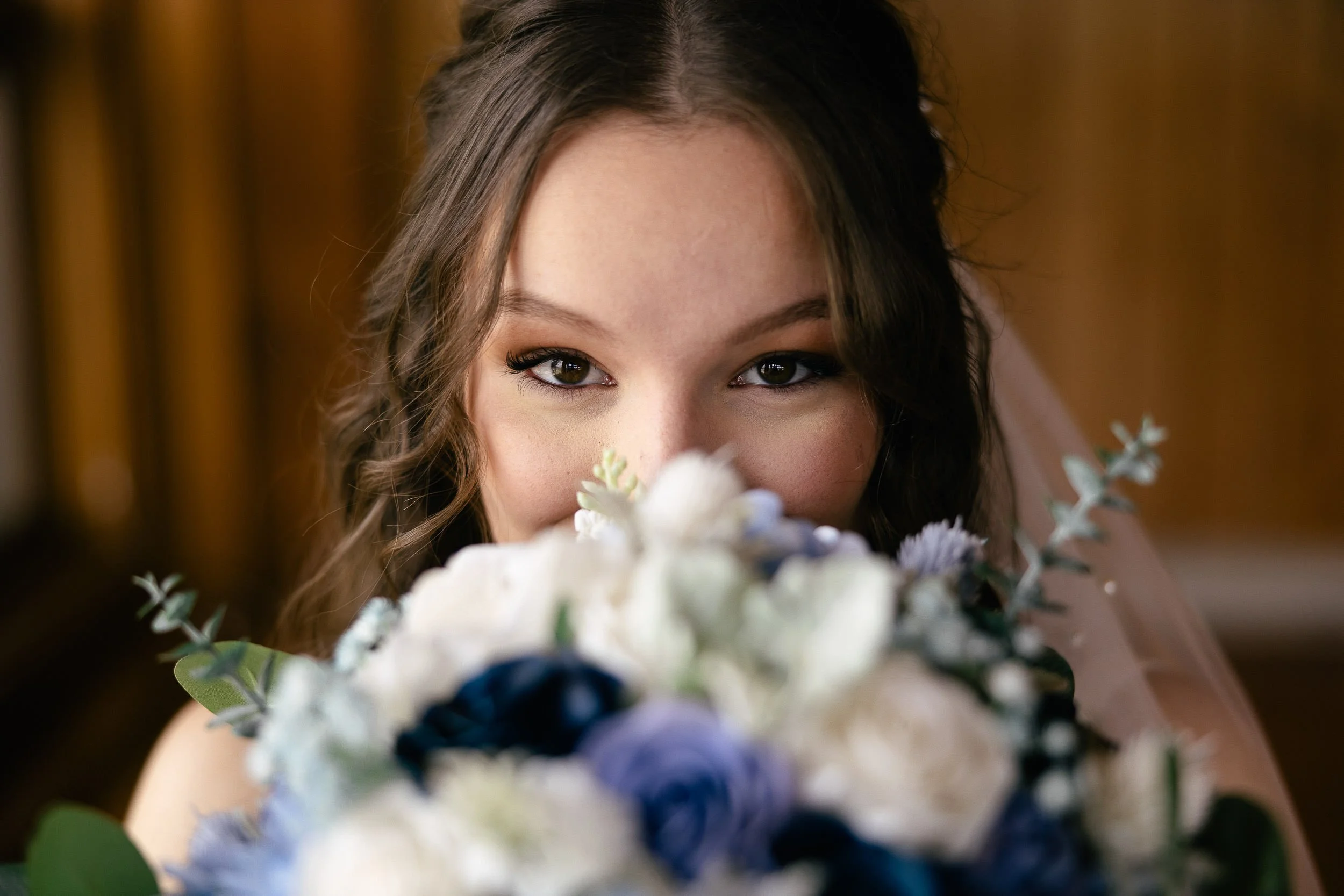 A woman with brown hair and brown eyes peeking over a wedding bouquet of white and purple flowers with green foliage, with a wooden background.