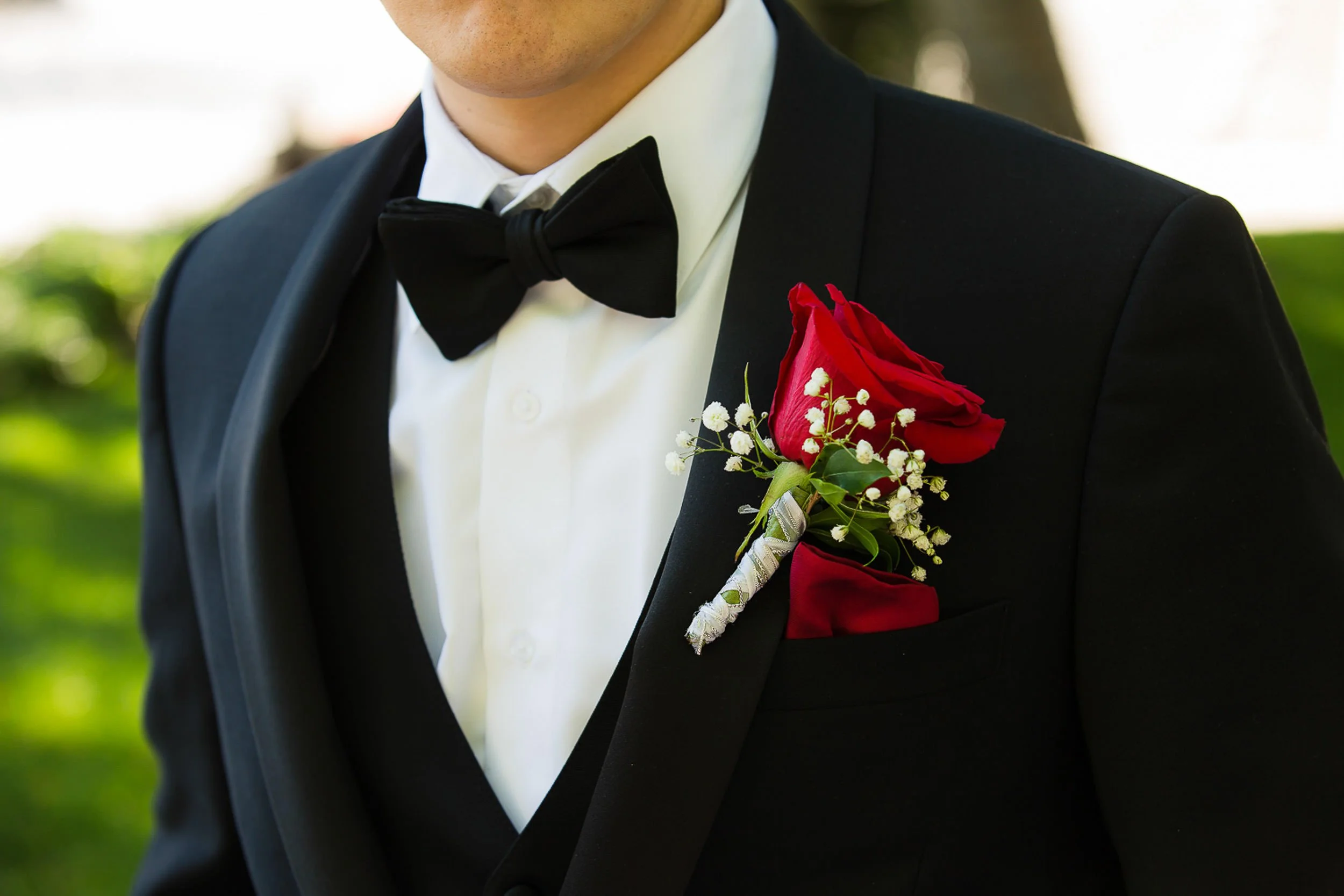 Close-up of a man in a tuxedo with a boutonniere of a red rose, white baby's breath, and greenery on his left lapel.