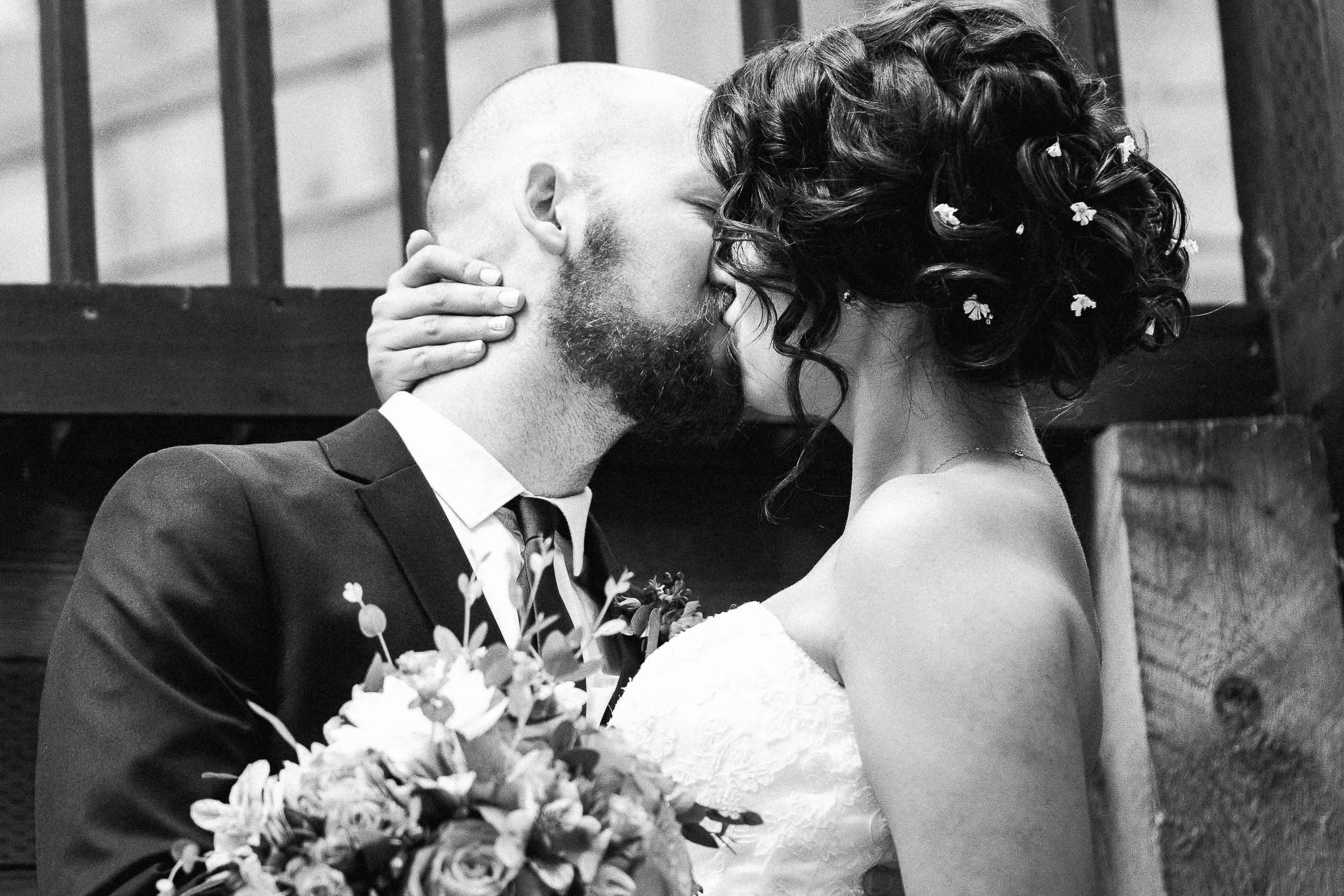 A black and white photo of a couple sharing a romantic kiss, with the woman holding a bouquet of flowers and dressed in a wedding gown, and the man dressed in a suit and tie.