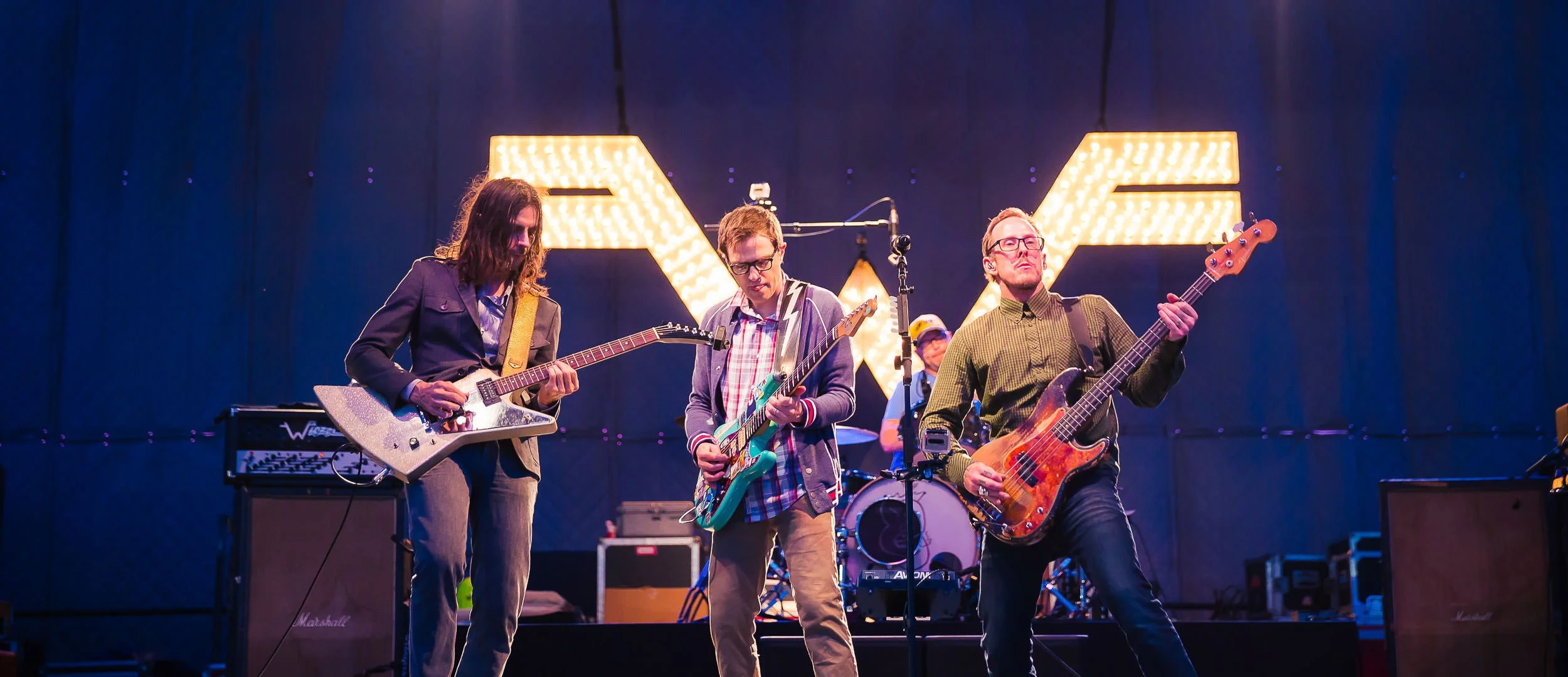 A band performing on stage with three guitarists in front and a drummer in the background. Bright illuminated letter signs are behind them.