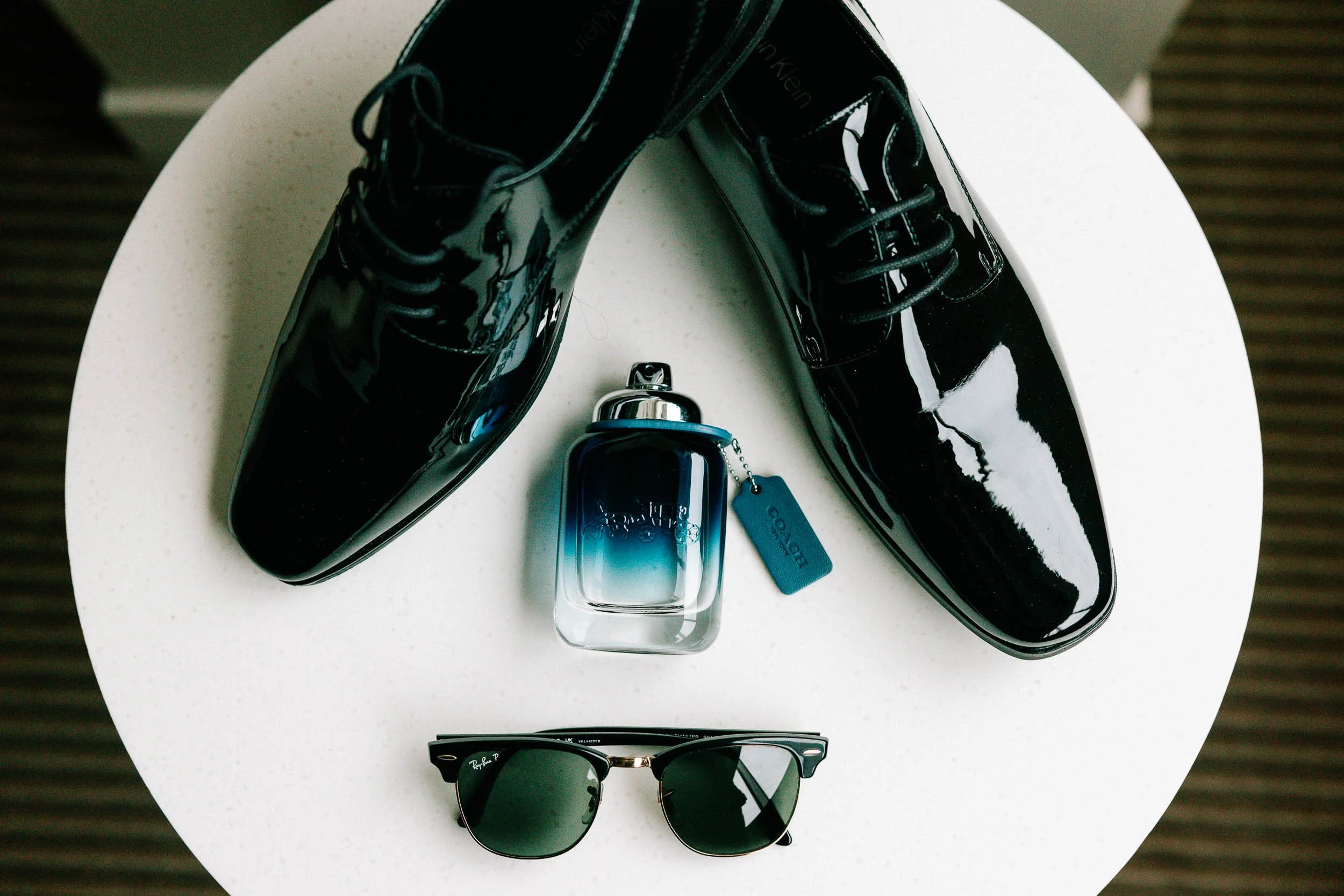 A pair of shiny black Calvin Klein boots, a small blue Loewe perfume bottle, a pair of black Ray-Ban sunglasses, and a pair of black sunglasses with gold accents on a white round table.