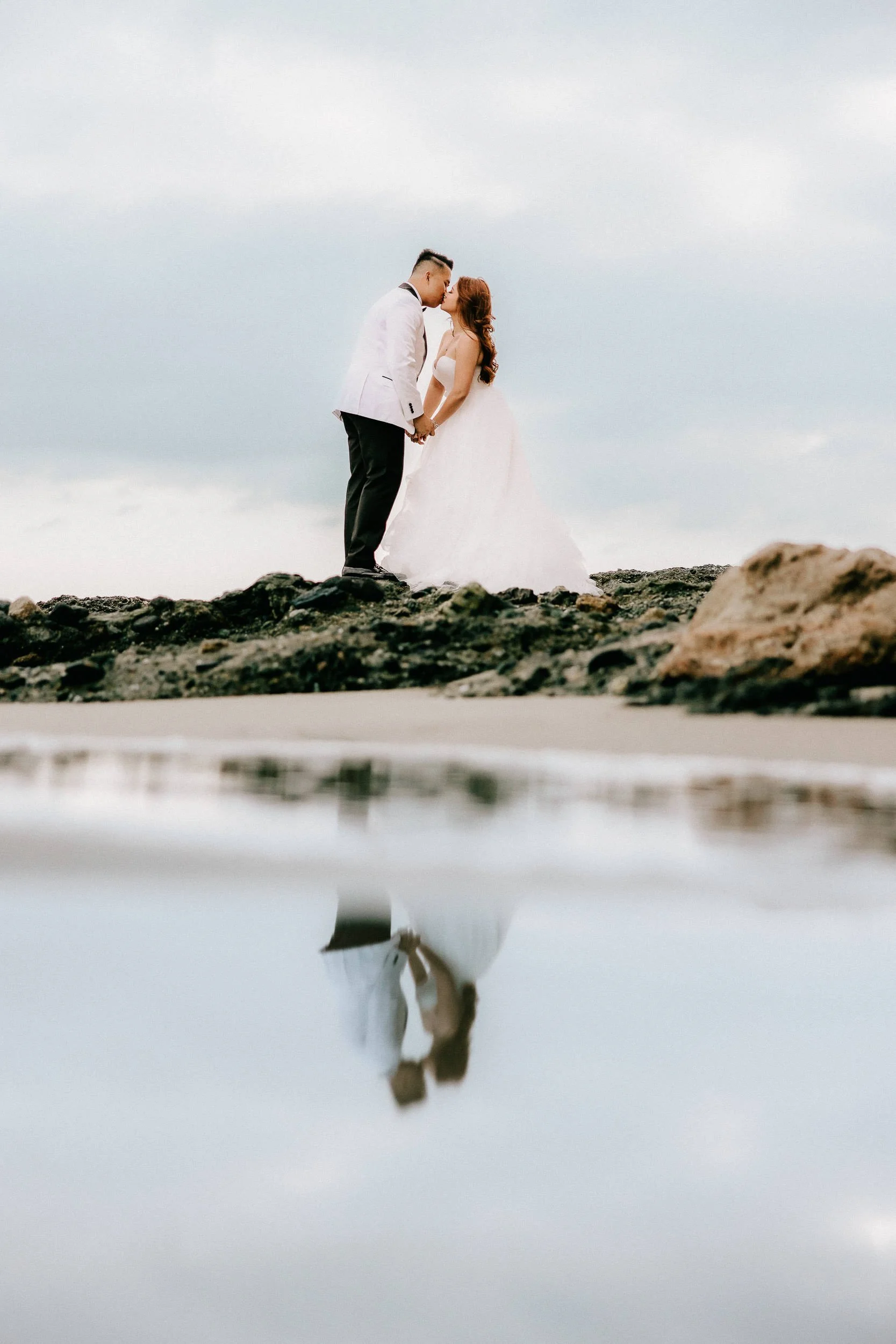 A bride and groom standing on rocks at the beach, sharing an intimate moment, with their reflection visible in the water below.