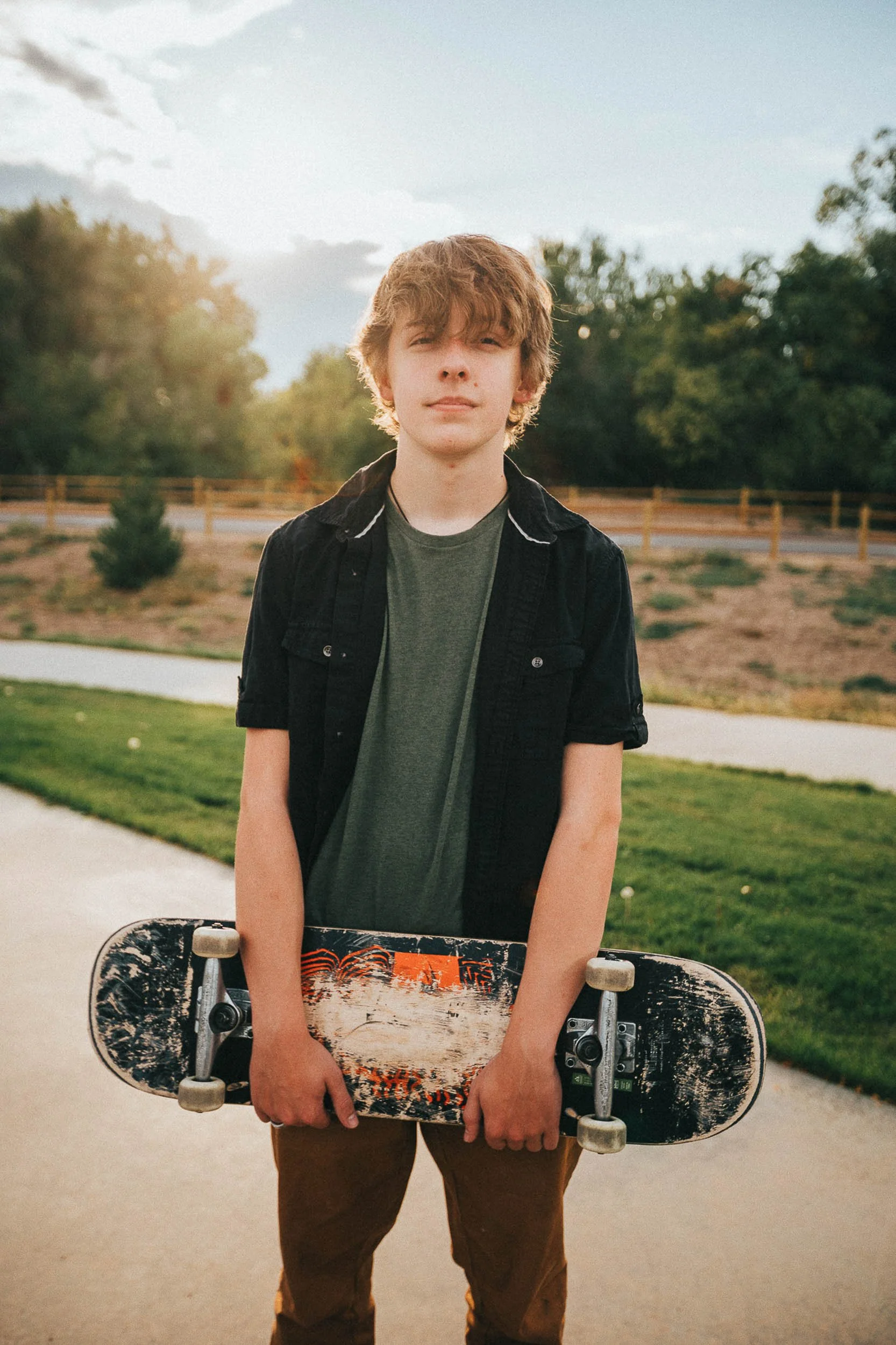 A young male teenager with curly, light brown hair holding a skateboard outdoors in a park-like setting with trees, grass, and a pathway, during daytime.