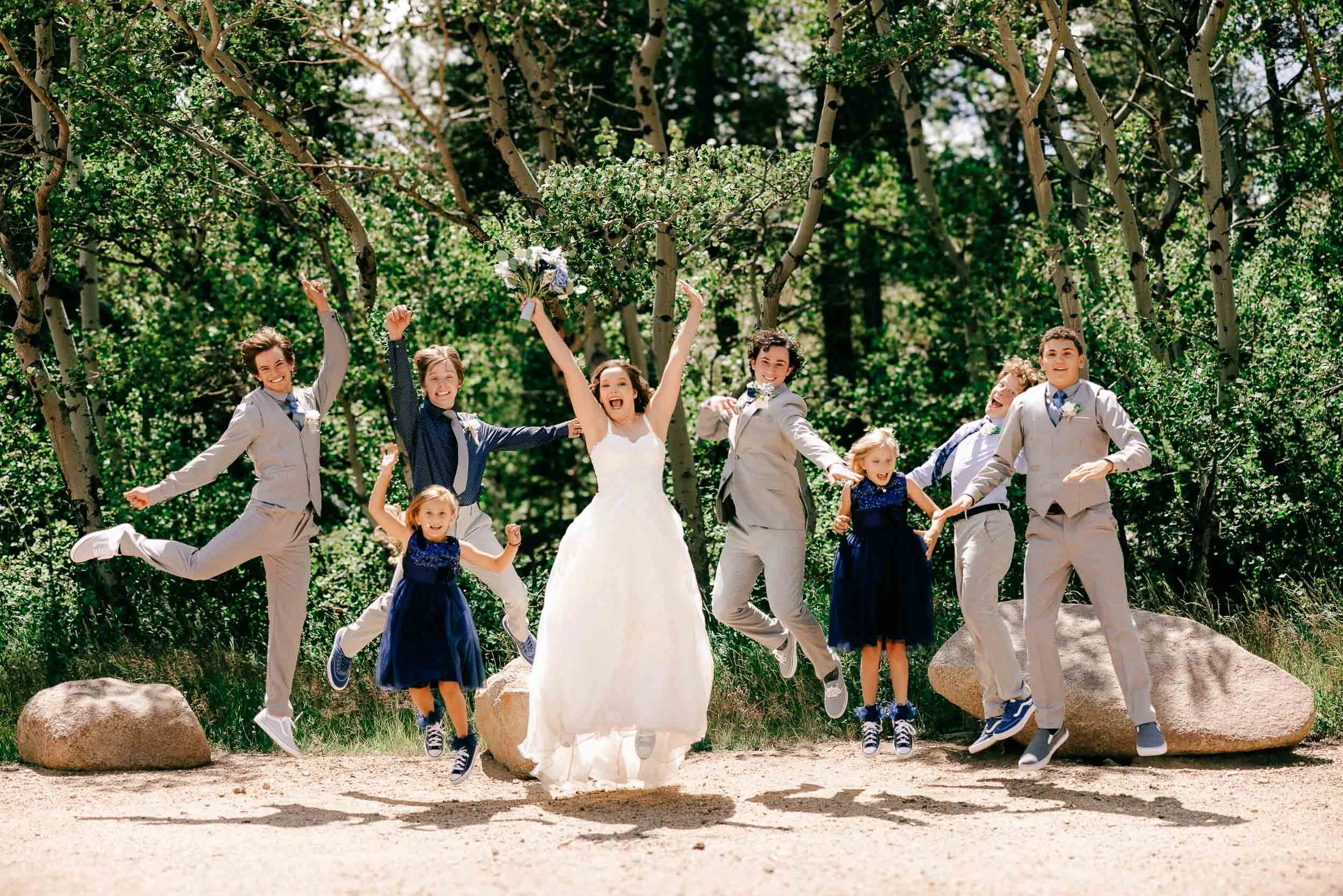 A group of people, including children and adults, celebrating outdoors in a wooded area during daytime. A woman in a white dress, a bride, is in the center holding a bouquet and jumping in joy. The others are dressed in formal attire, most of them ju
