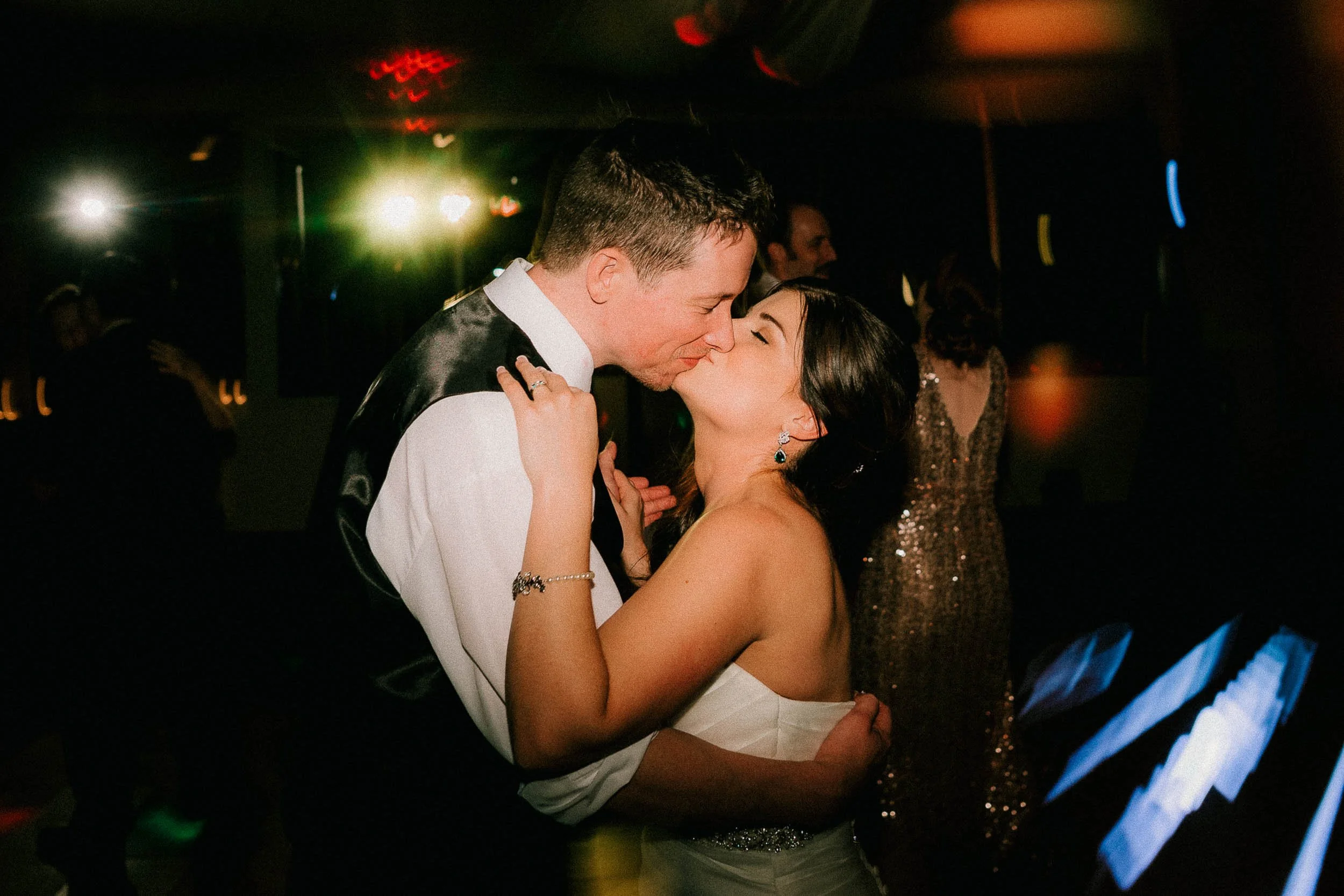 A couple is dancing closely at a party or wedding reception in a dimly lit room, with colorful lights in the background.