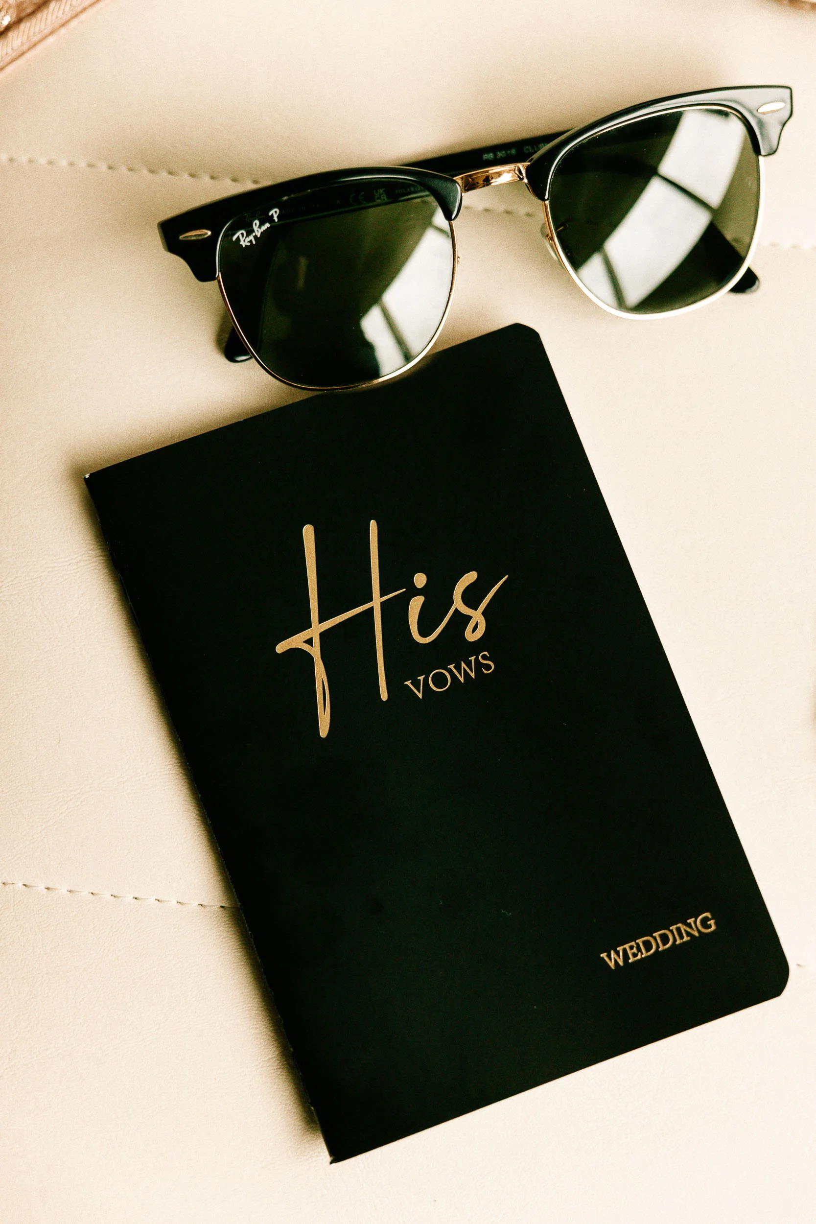 Black and gold sunglasses resting on a cream-colored surface, with a black box labeled 'His Vows Wedding' partially visible underneath.