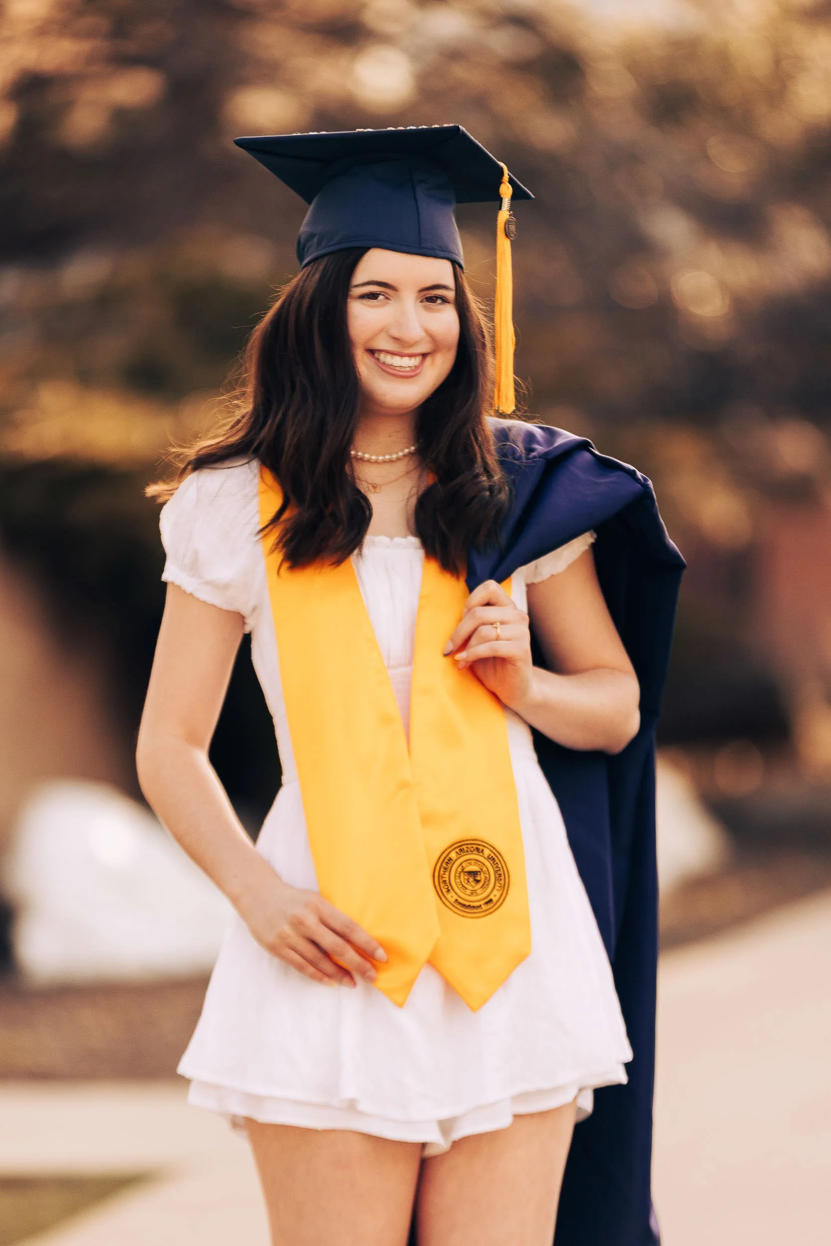 Young woman in white dress holding a yellow graduation stole with official emblem, wearing a blue graduation cap and gown, celebration outdoors.