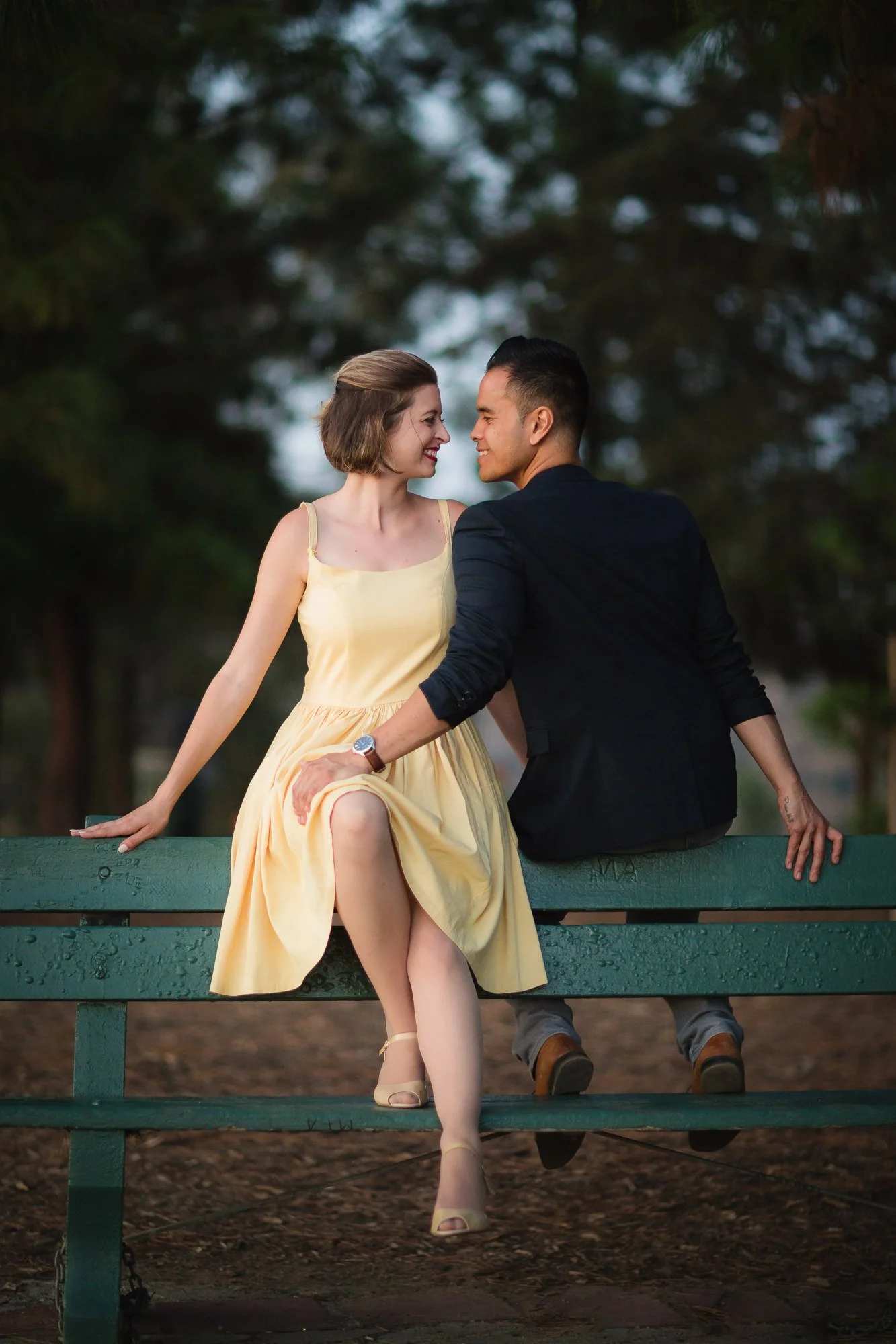 A woman in a yellow dress and heels sitting on a green bench with a man in a black jacket and gray pants, both smiling and touching noses, outdoors in a park with trees in the background.
