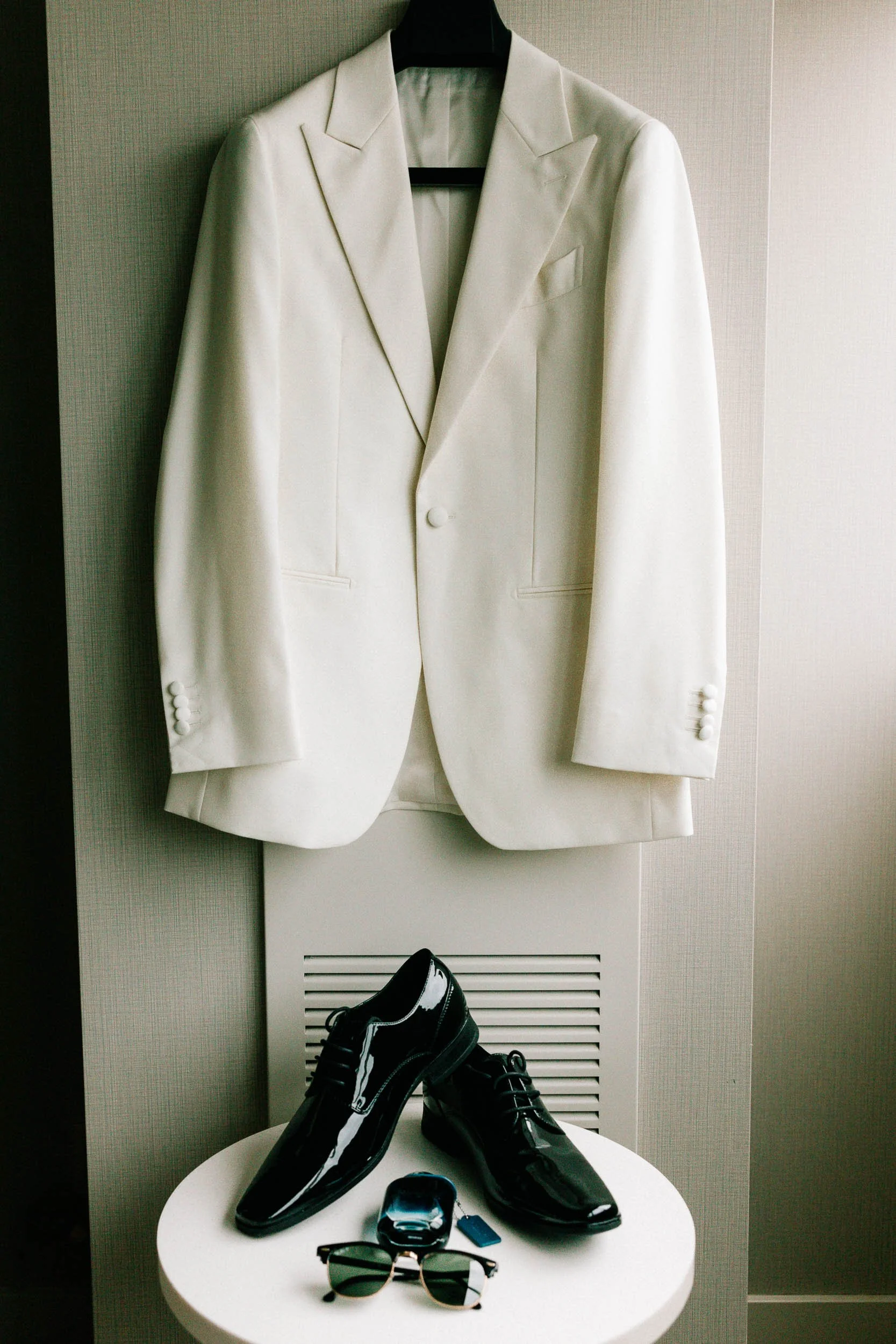 White blazer hanging on a black hanger on a door, black dress shoes on a small white table, sunglasses, a fitbit, and a keychain on the table.