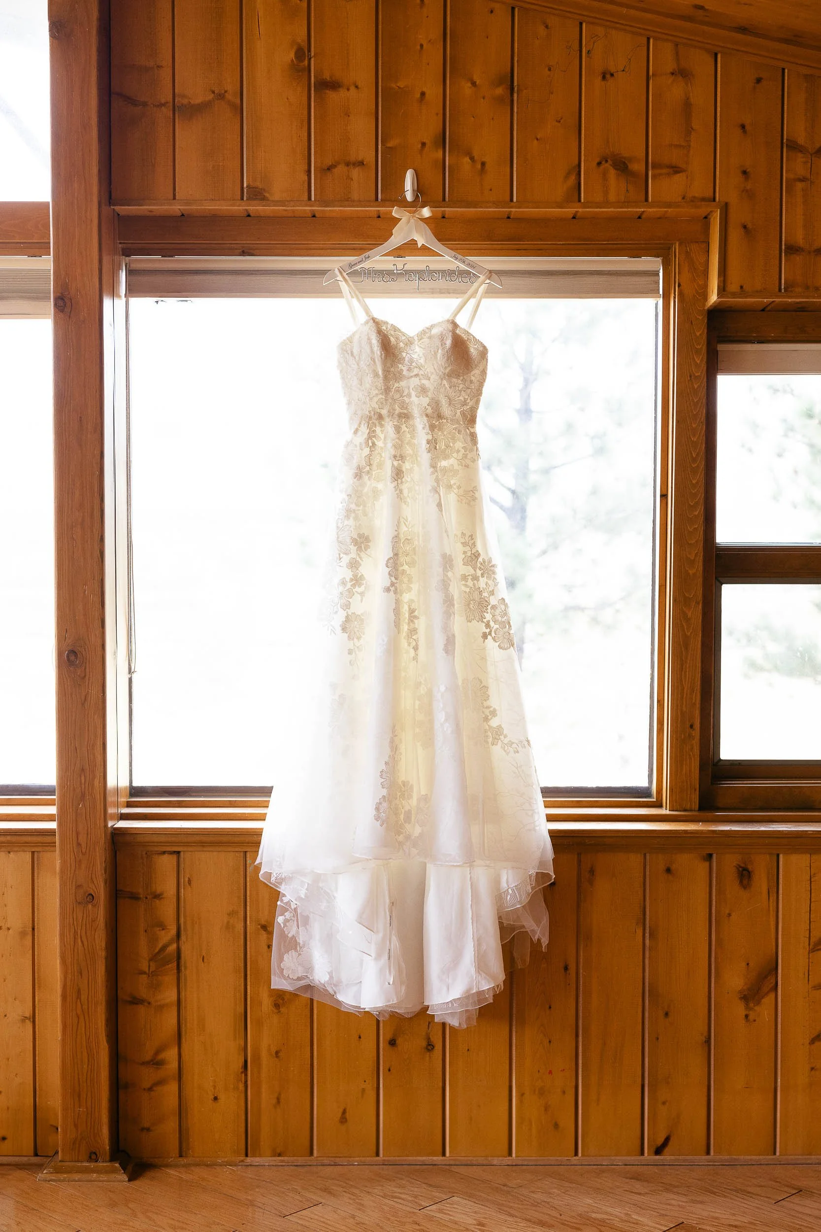 A white wedding dress with floral lace embroidery hanging from a wooden beam in front of a large window with a scenic view.