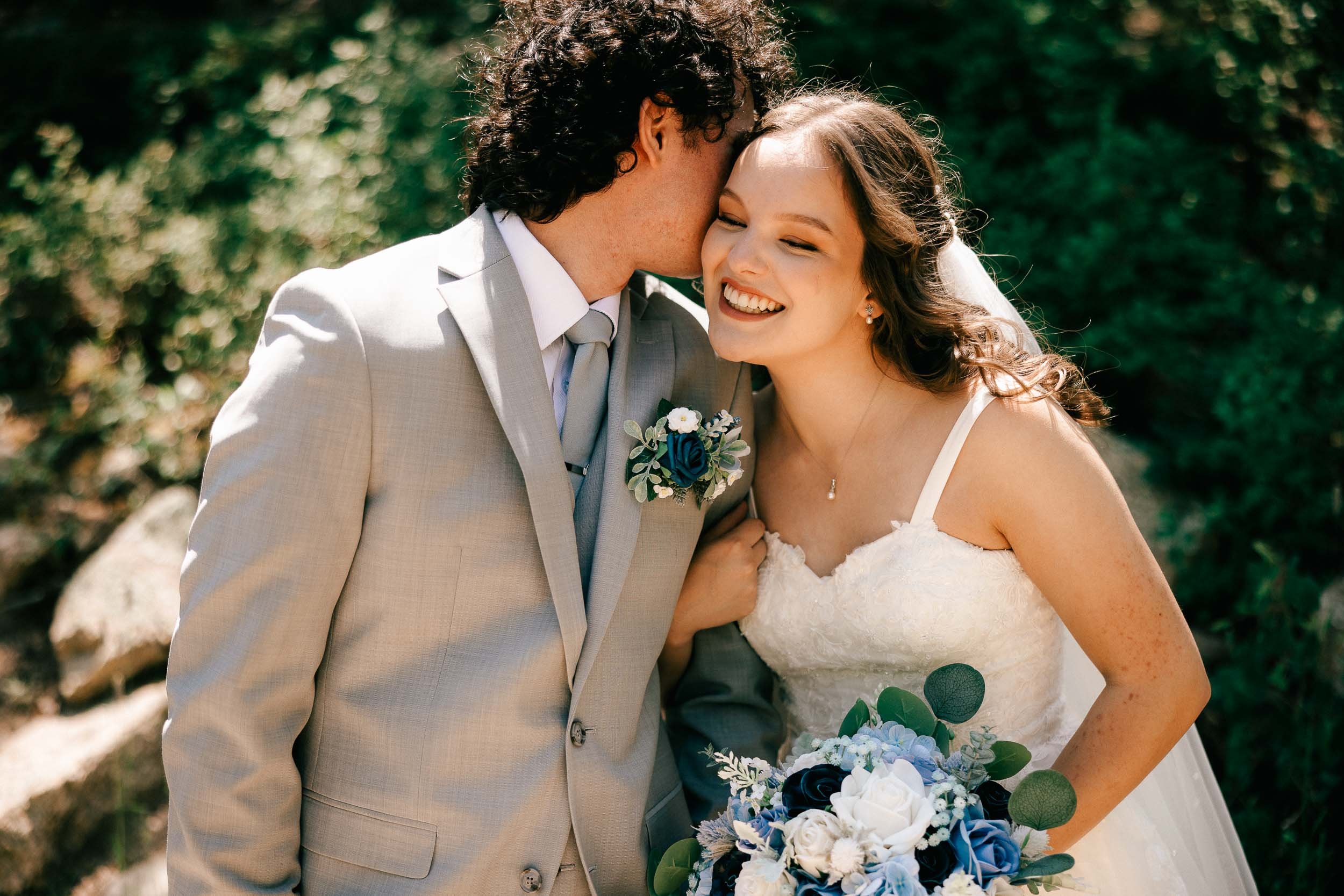 A bride and groom sharing a kiss outdoors, with the bride smiling and holding a bouquet of white and blue flowers, and the groom wearing a light gray suit with a boutonnière, against a blurred green background.