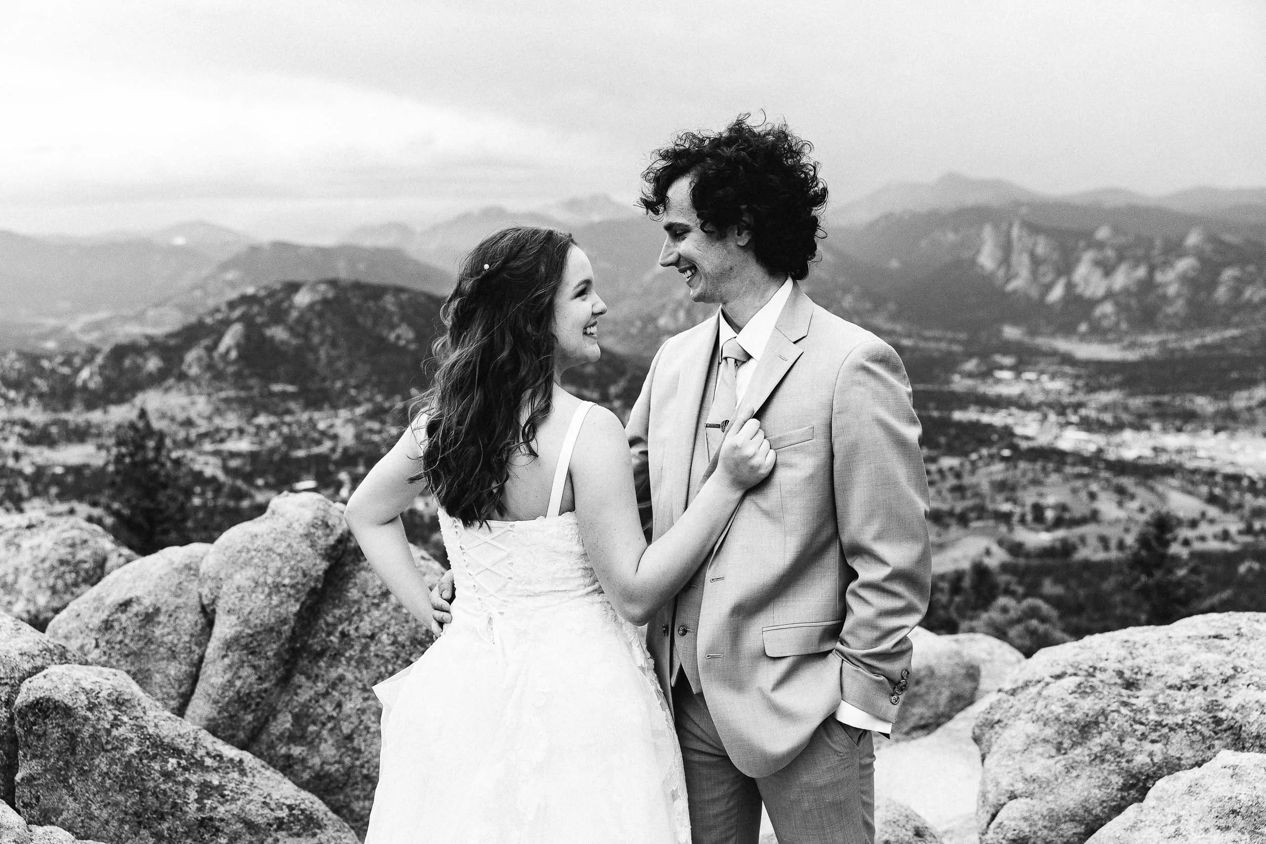 A black and white photo of a bride and groom standing on rocks outdoors with mountains in the background, smiling at each other.