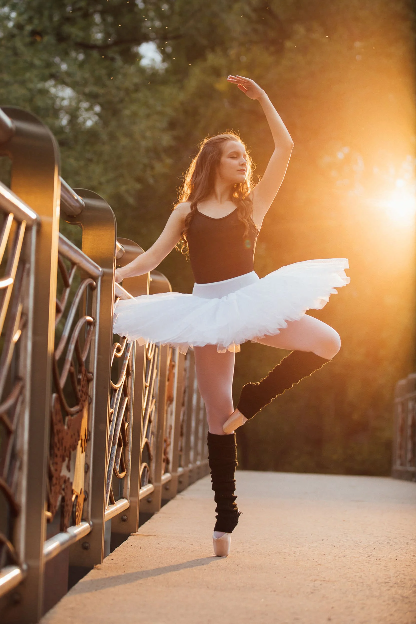 Ballet dancer standing on one leg in outdoor setting during sunset, wearing a black top, white tutu, and black leg warmers, with sunlight behind her.
