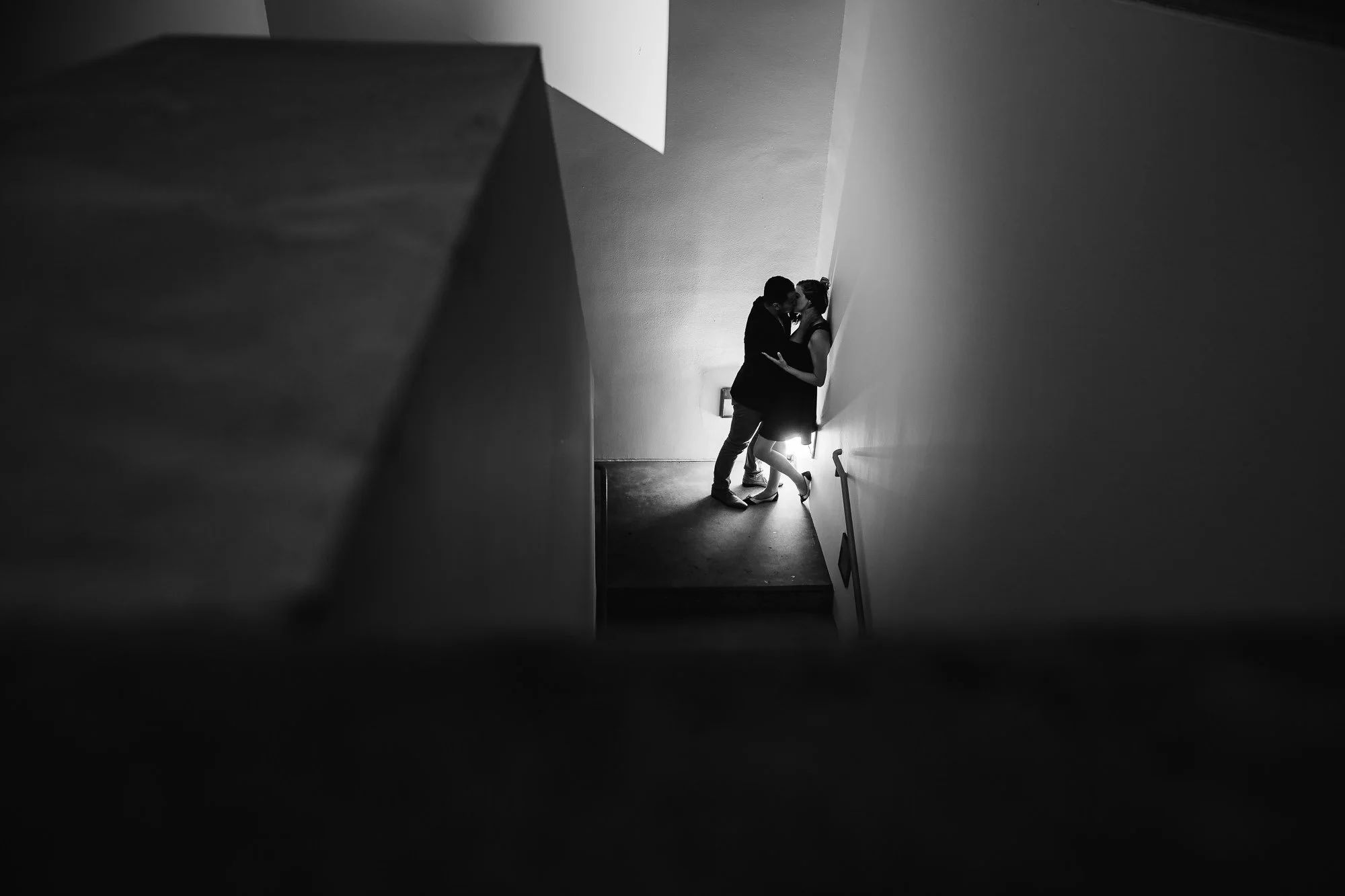 A black and white photo of a couple kissing in a stairwell. The image is viewed from above, with the couple standing against a plain wall near a small window.